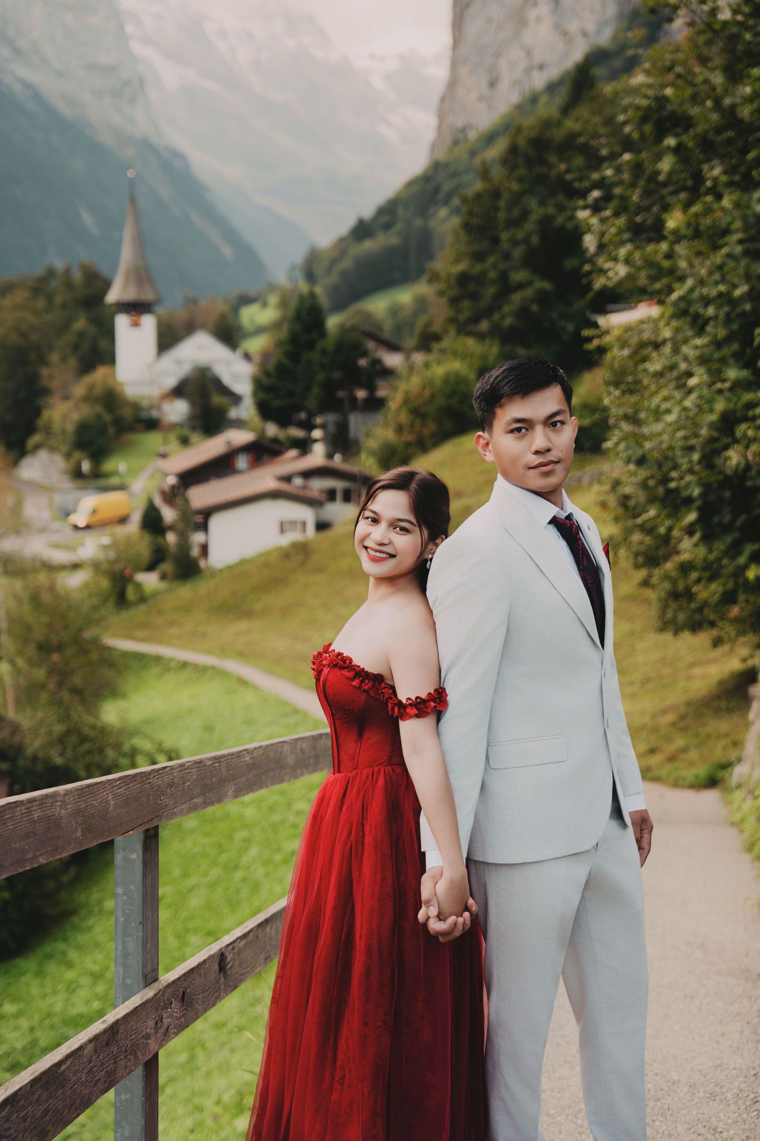 Elegant couple holding hands on scenic Swiss alpine path, woman in flowing red gown smiling.