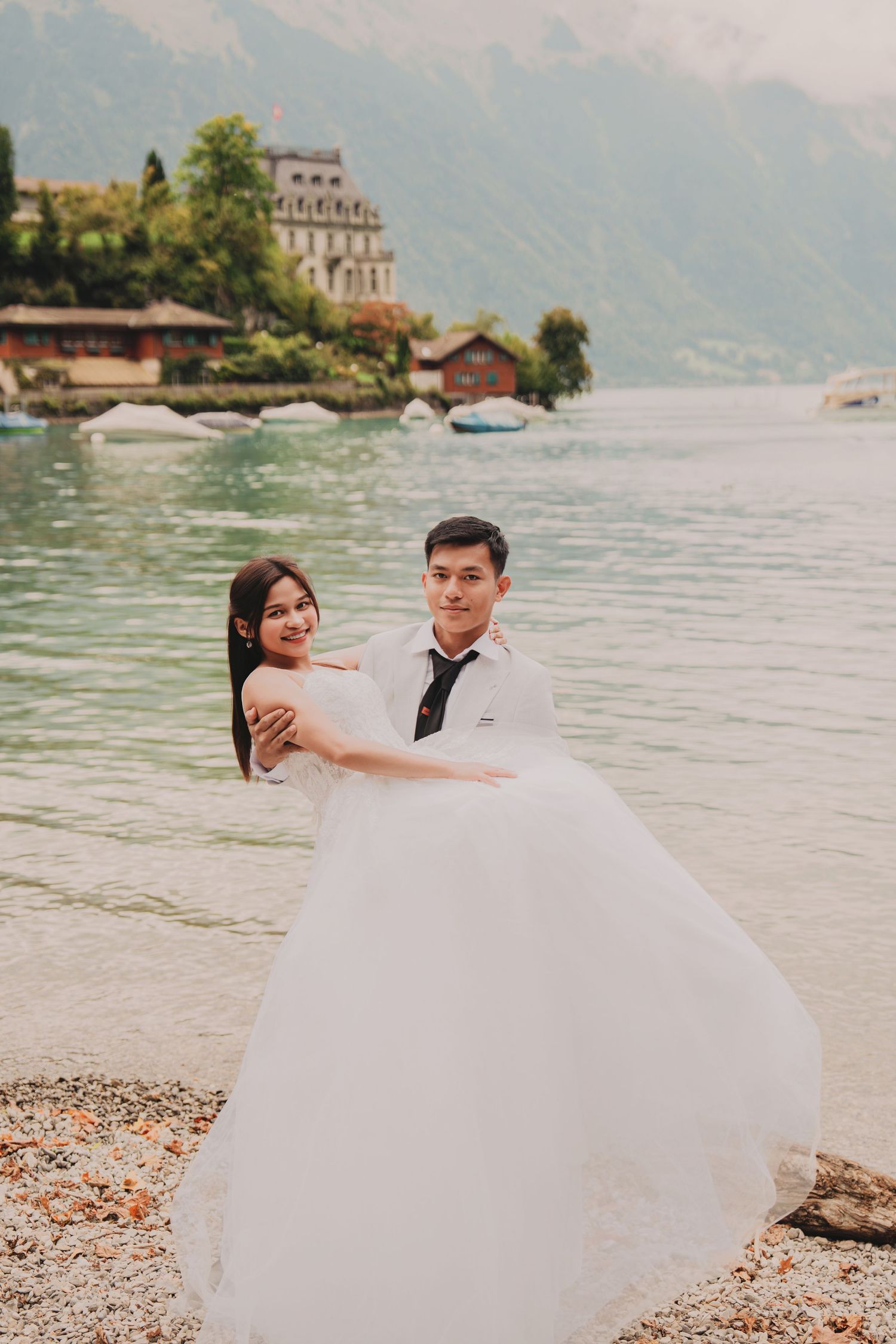Groom lifting bride in flowing white dress on rocky lakeshore with alpine scenery and château backdrop.