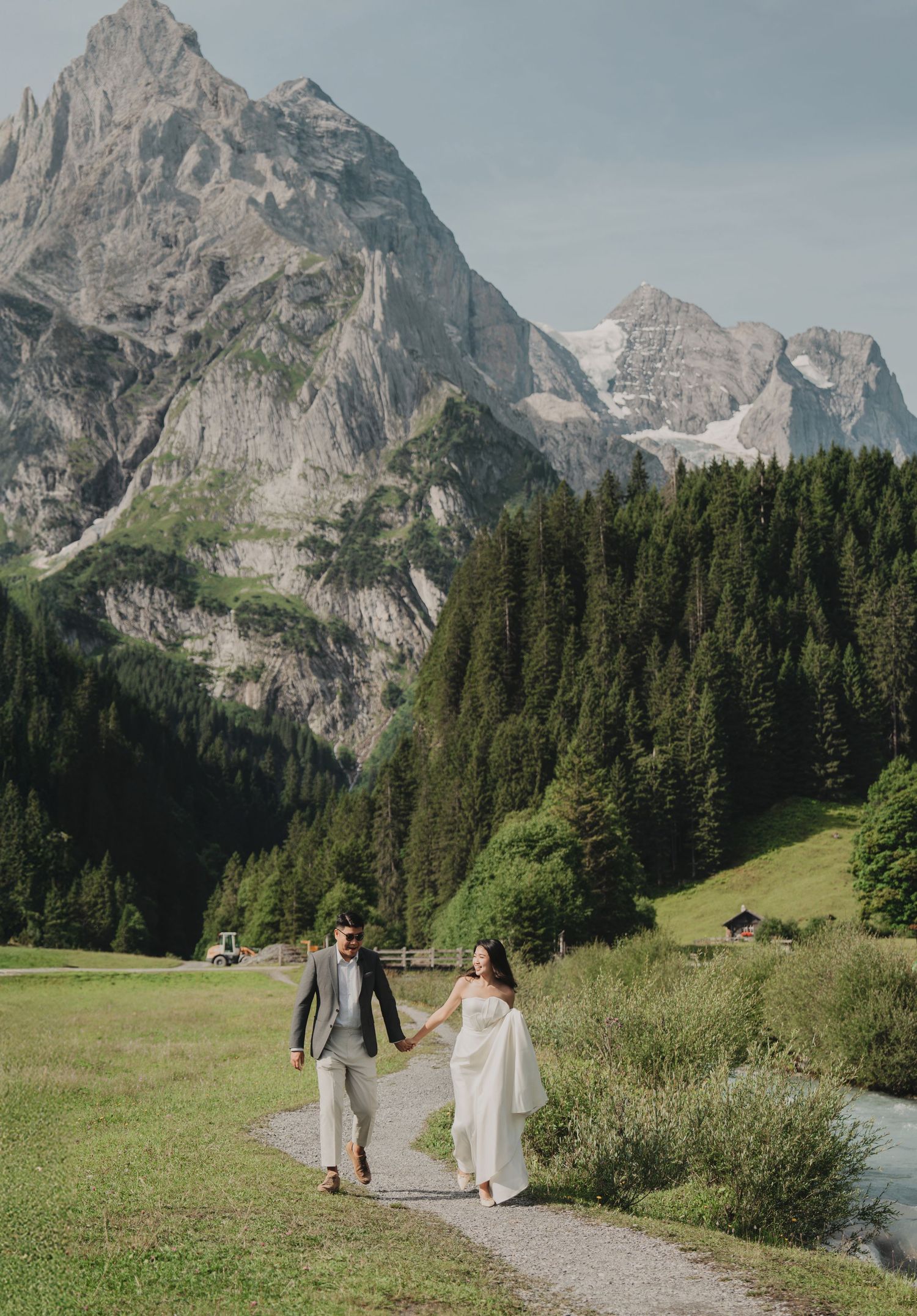 Couple holding hands on a mountain trail beside a stream with stunning rocky peaks and green forest behind them.
