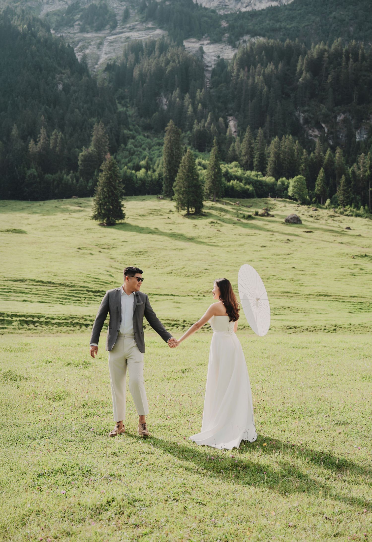 Wedding couple holds hands in lush green mountain valley, bride carrying white parasol on sunny day.