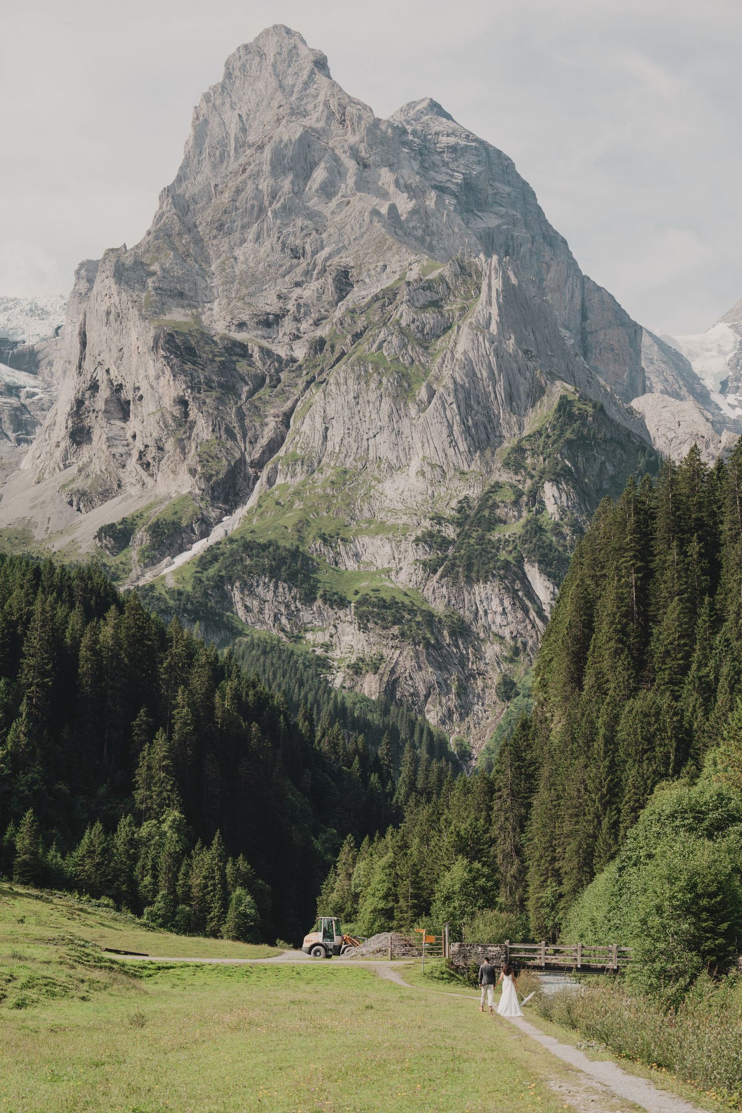 Tiny bride and groom figures walking a valley path dwarfed by a massive dramatic Alpine mountain peak.