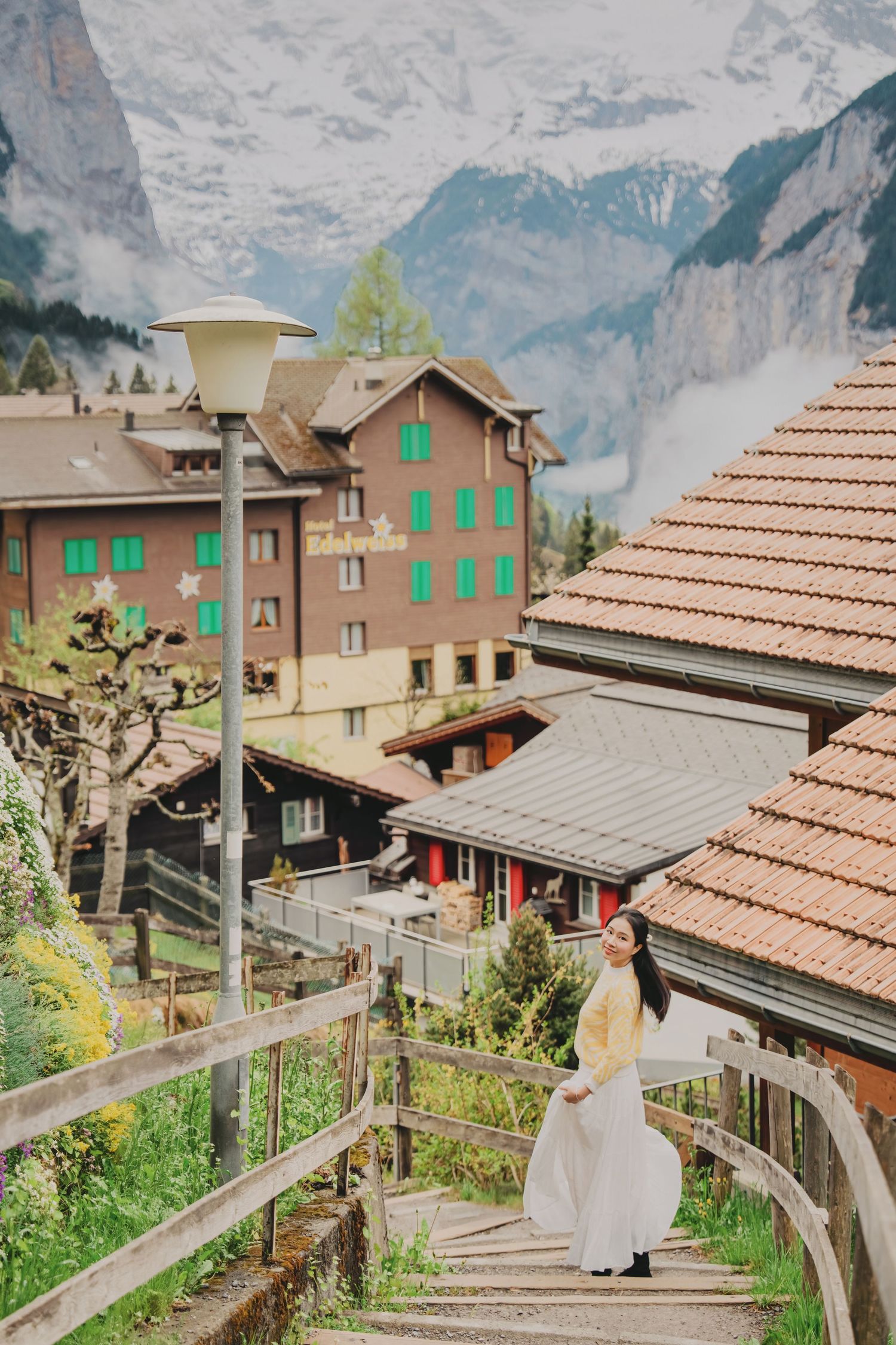 Woman in white skirt poses on wooden stairs with snow-capped Swiss Alps behind her.