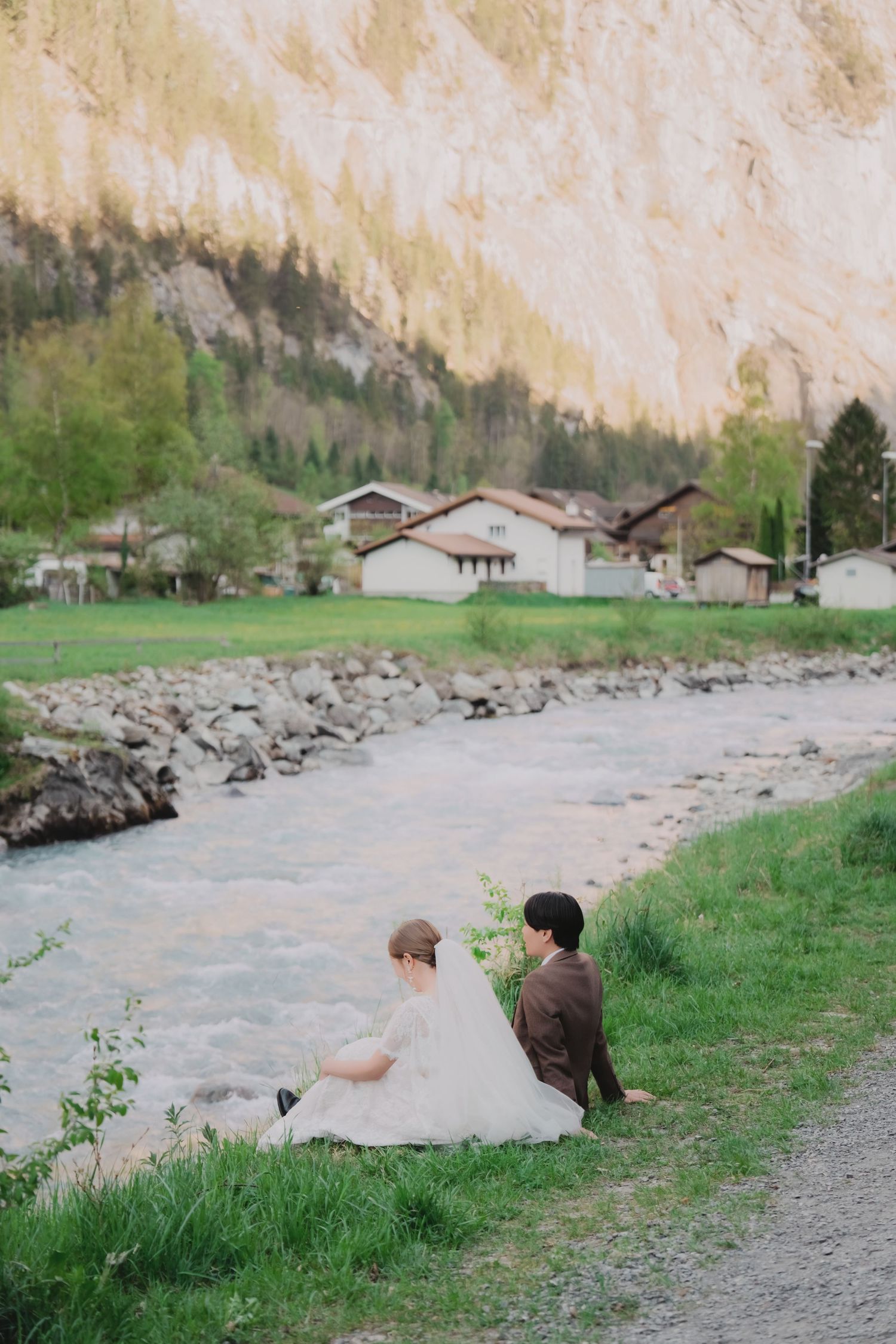 Wedding couple seated on grassy riverbank with dramatic Swiss mountain cliffs and alpine village in background.