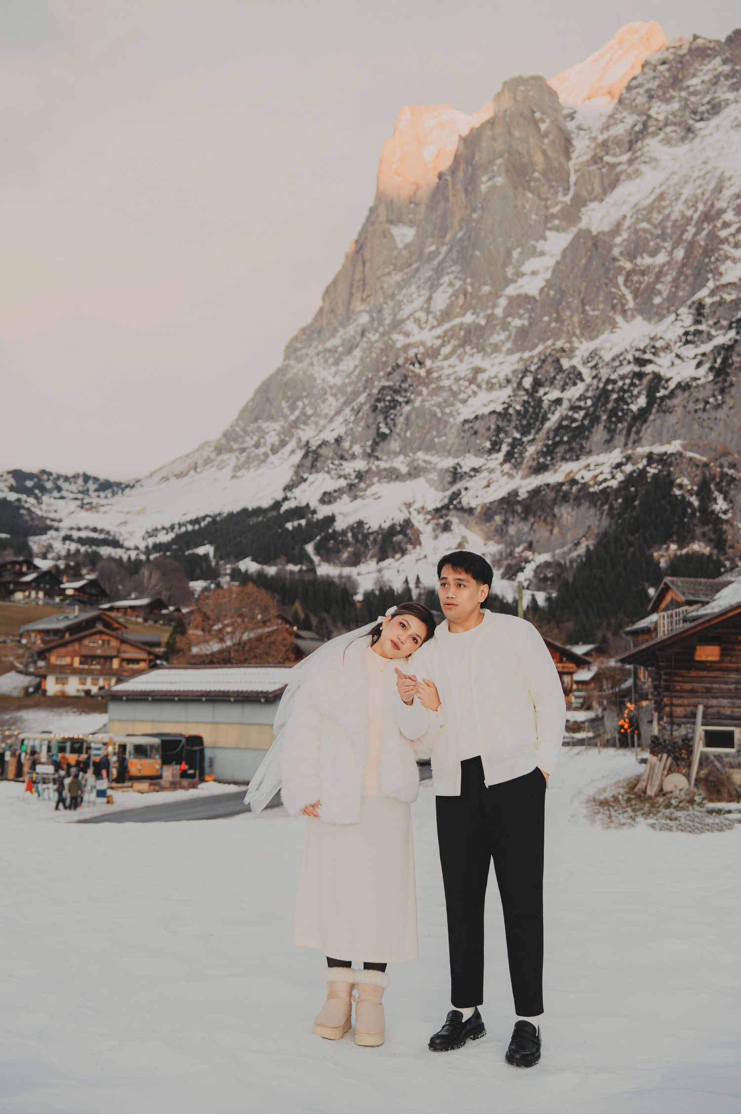 Couple posing in white outfits on snowy ground with alpine village and mountain backdrop at dusk.