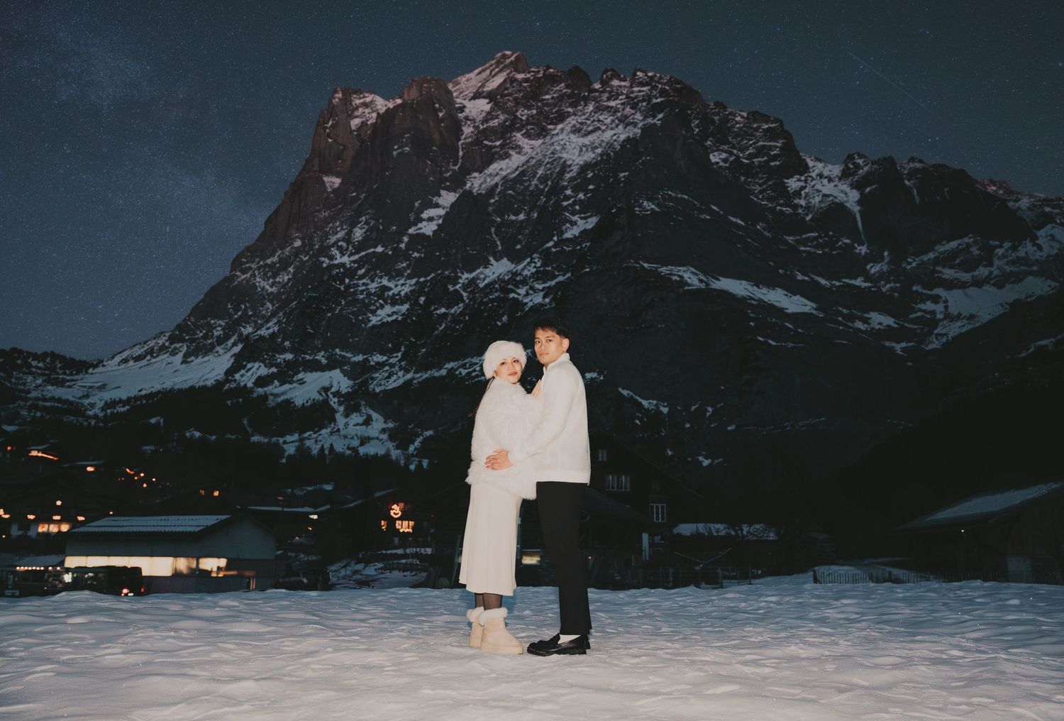 Couple posing in white winter attire before dramatic snow-capped mountain at night.