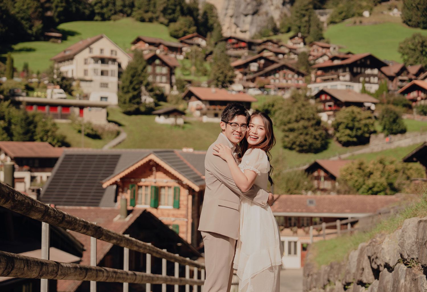 Couple hugging warmly with colorful Swiss alpine chalets and lush green hillside in the background.