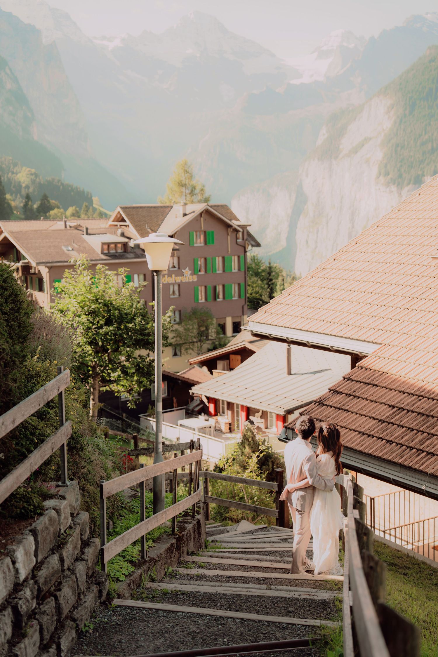 Couple descending wooden staircase in picturesque Swiss mountain village with misty alpine valley backdrop.