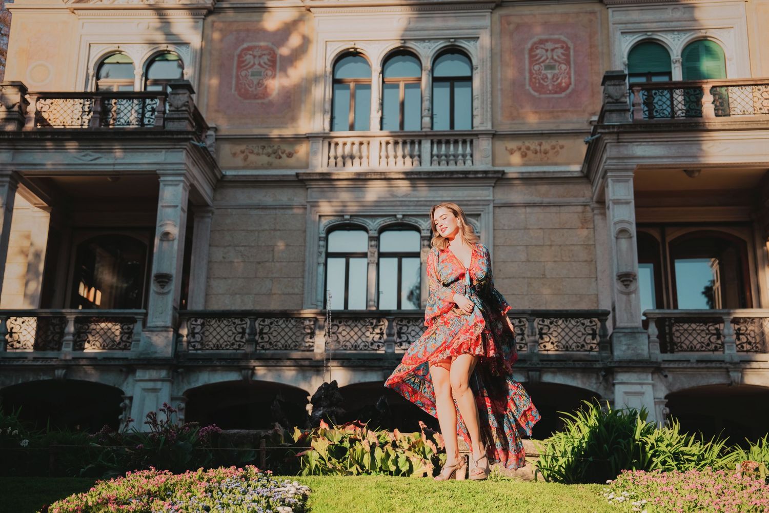 Woman in red floral dress stands before an ornate European-style mansion facade.