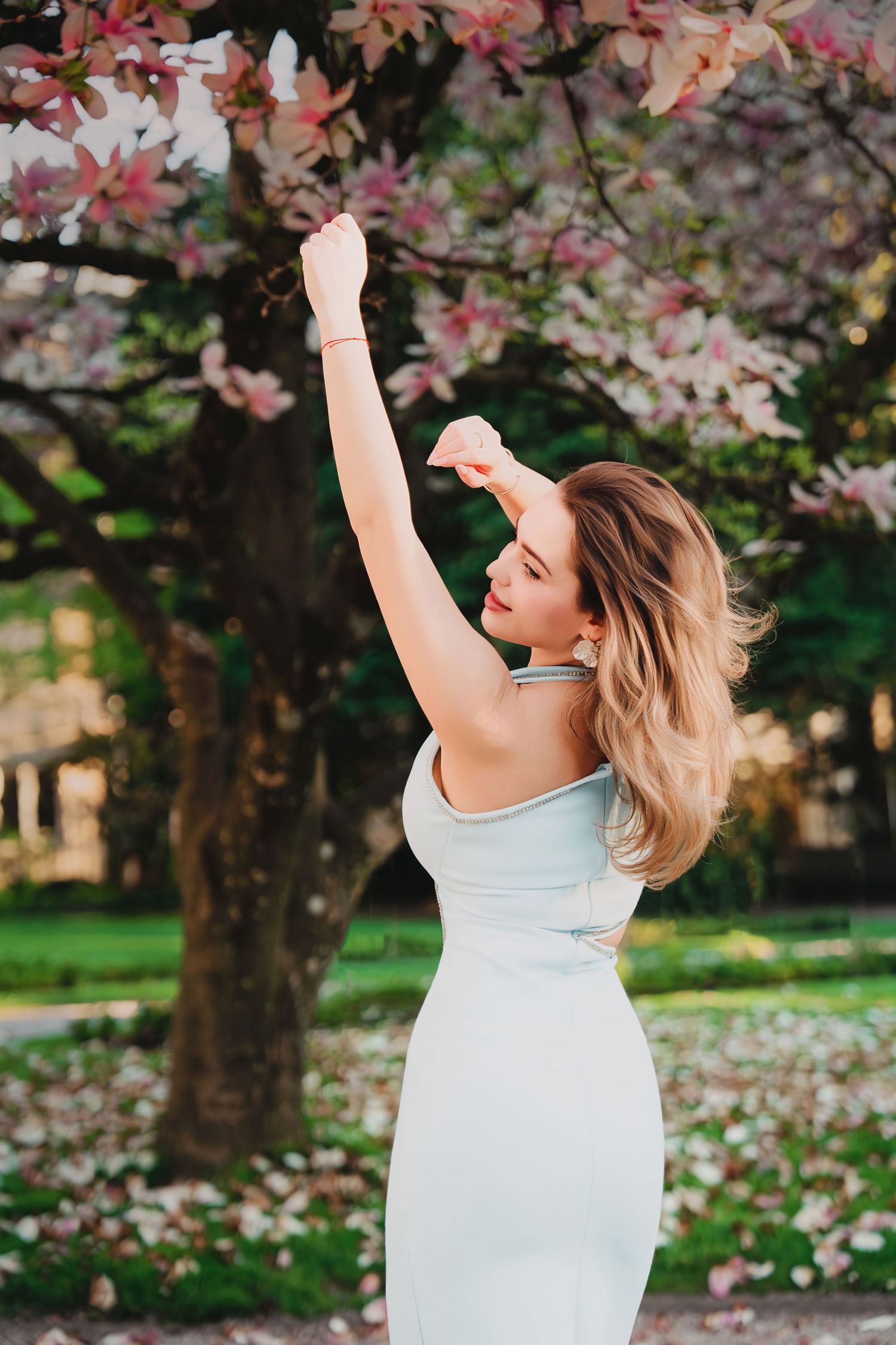Blonde woman in white dress reaching up to touch pink magnolia blossoms in garden.