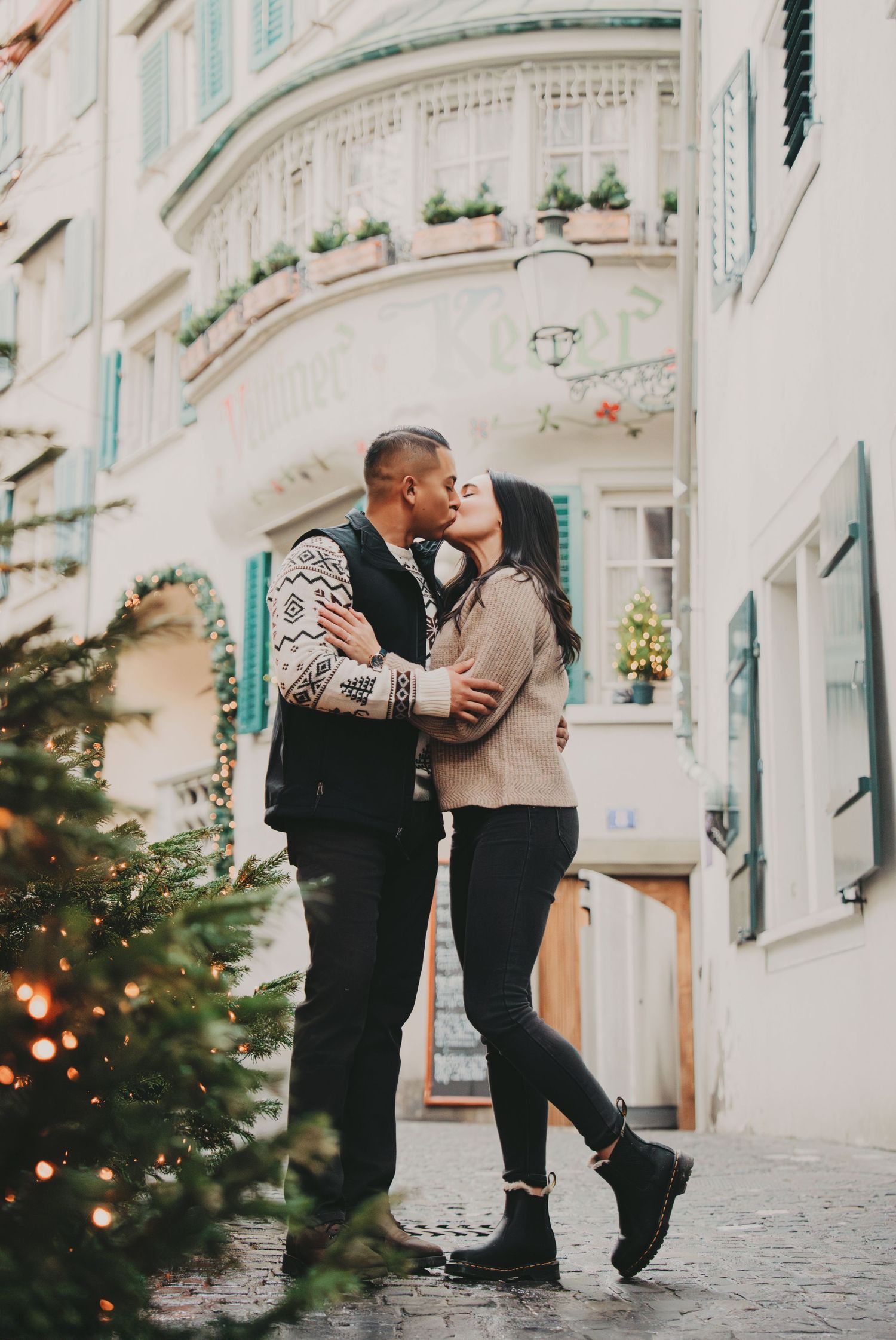 Couple sharing a romantic kiss near a Christmas tree on a charming European cobblestone street.