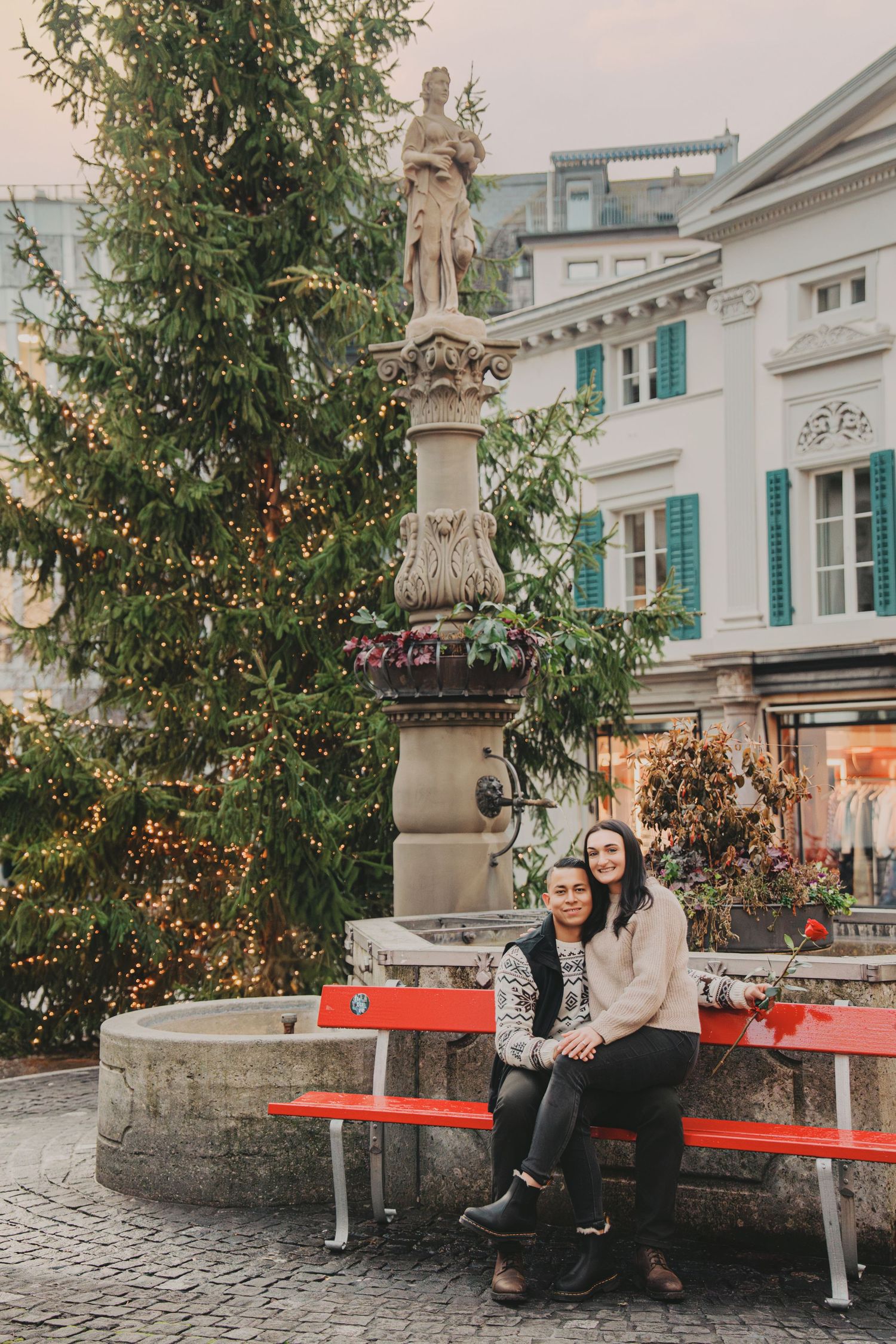 Couple sitting on red bench near fountain and Christmas tree in European square.