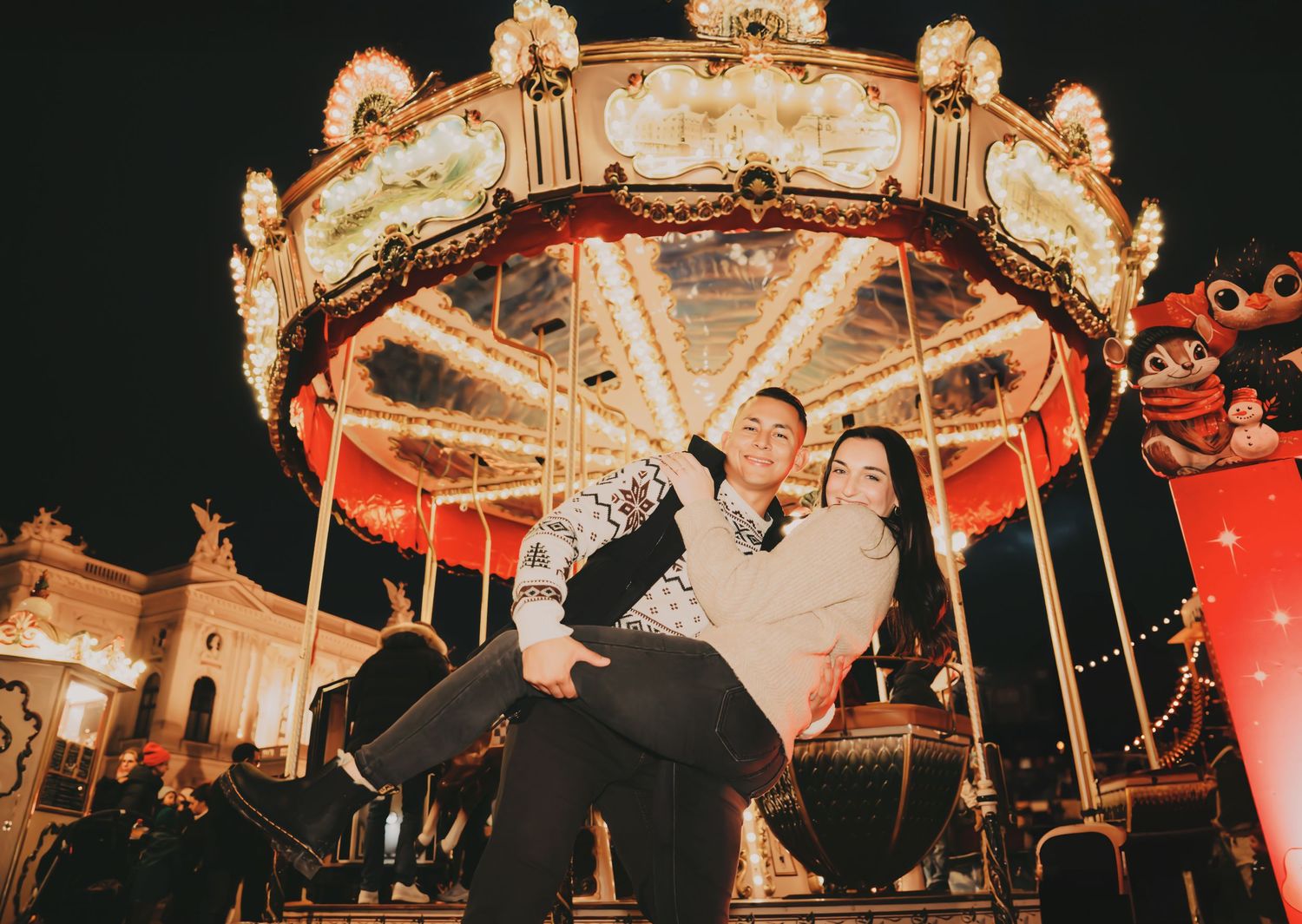 Man playfully lifting woman in front of a brightly lit vintage carousel at a Christmas night fair.