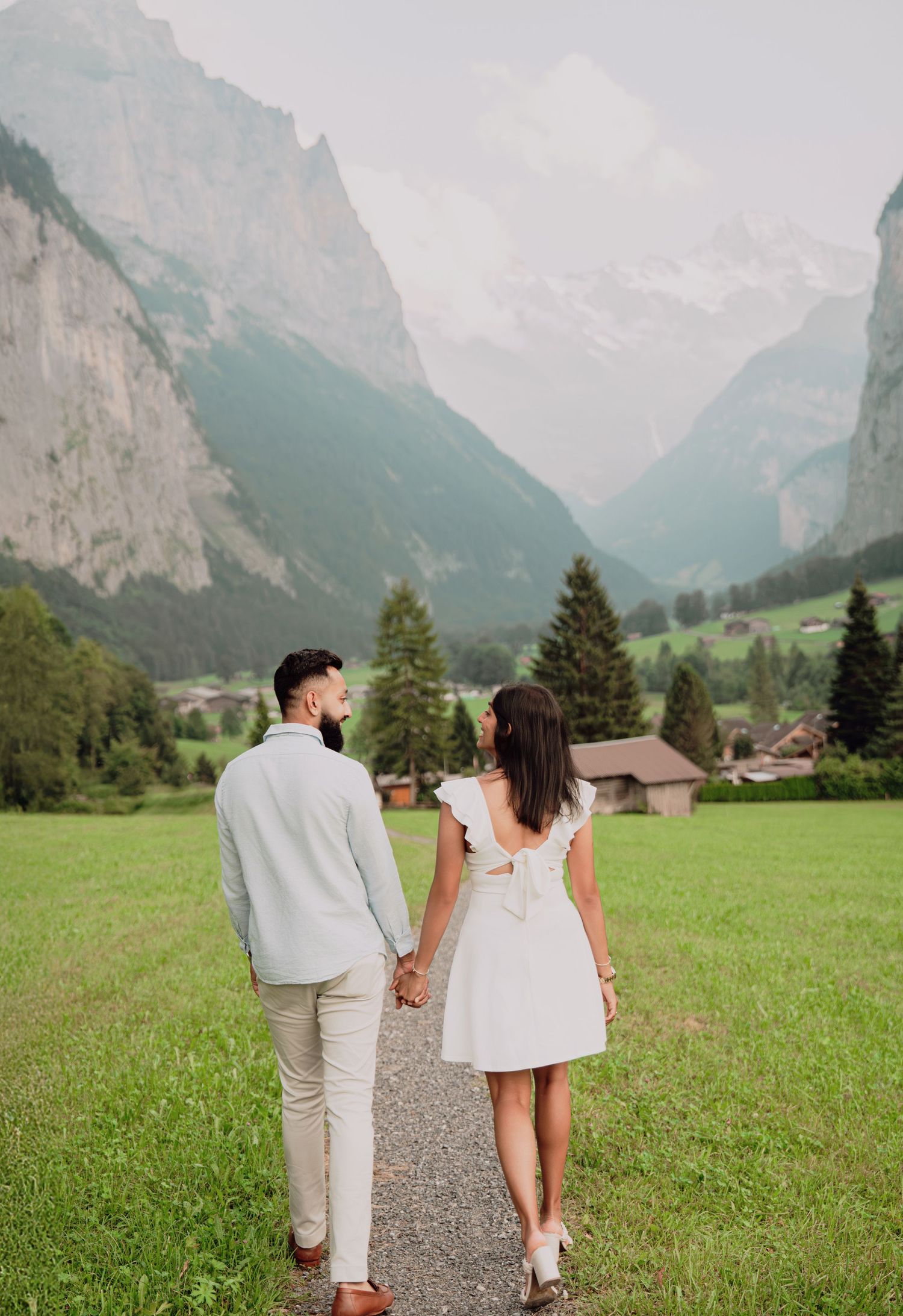 Couple walking hand in hand on a path through a lush green Swiss valley with majestic snow-capped mountains.