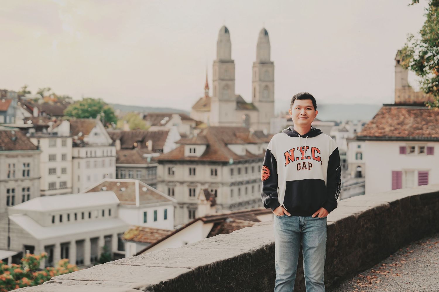 Young Asian man in NYC Gap hoodie standing by stone wall with Zurich cityscape behind him.