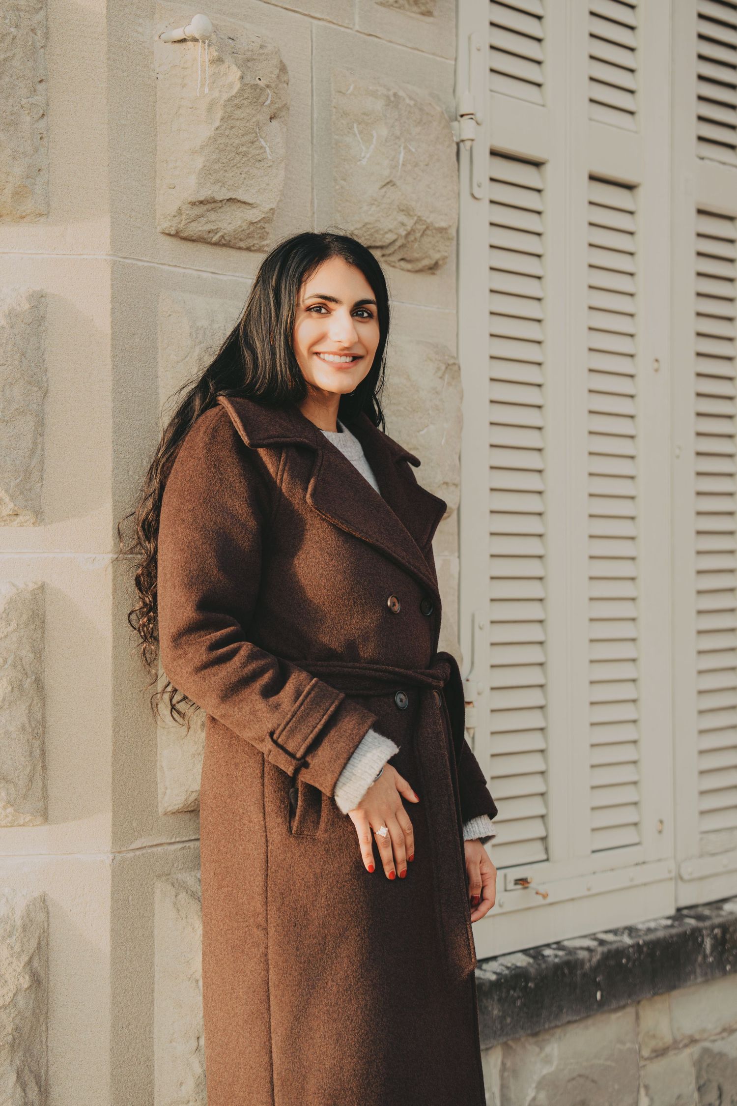 Happy woman in brown belted coat smiling while leaning against a stone wall with shutters.