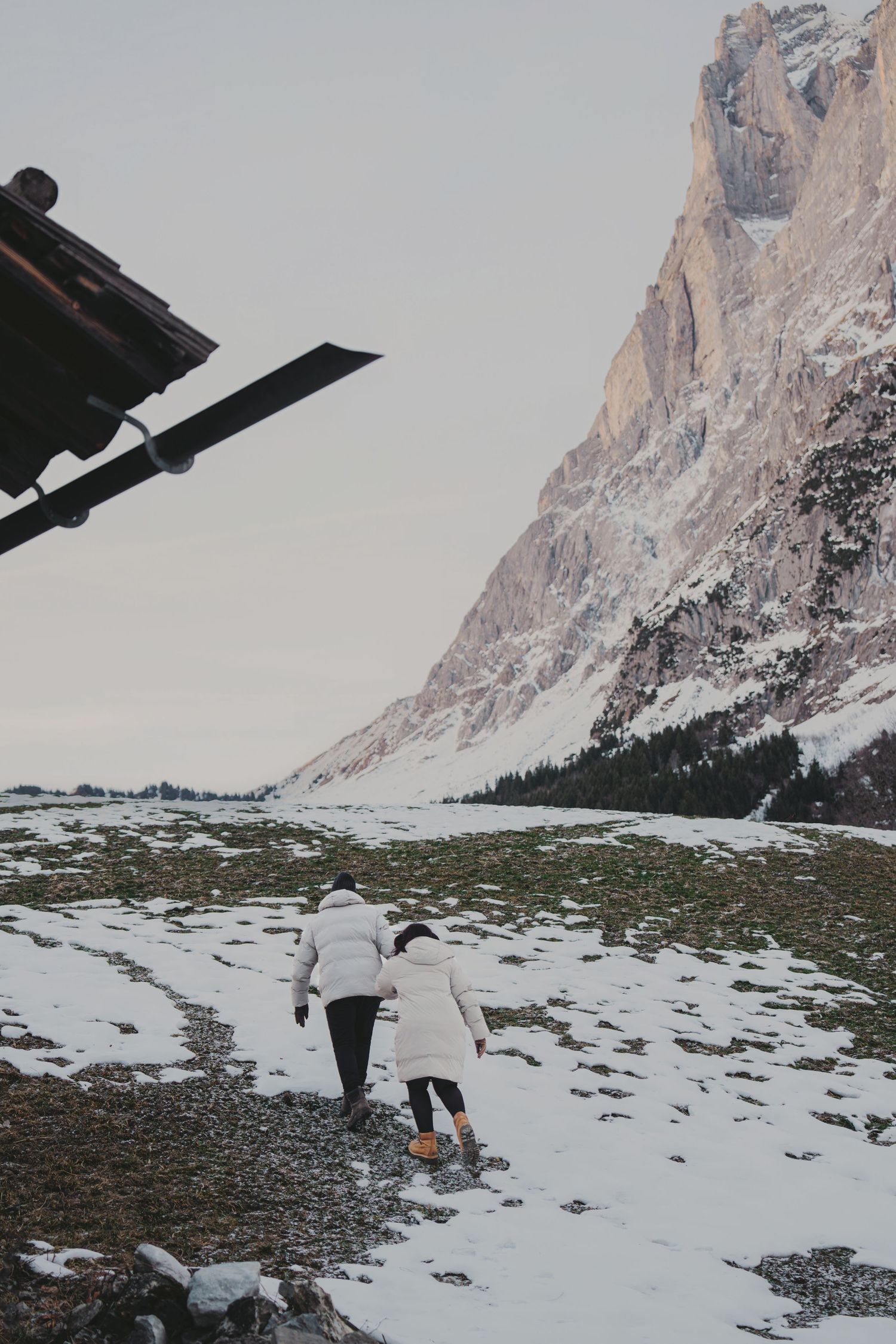 Two people in white jackets walking toward snow-covered valley beneath towering rocky mountain cliffs.