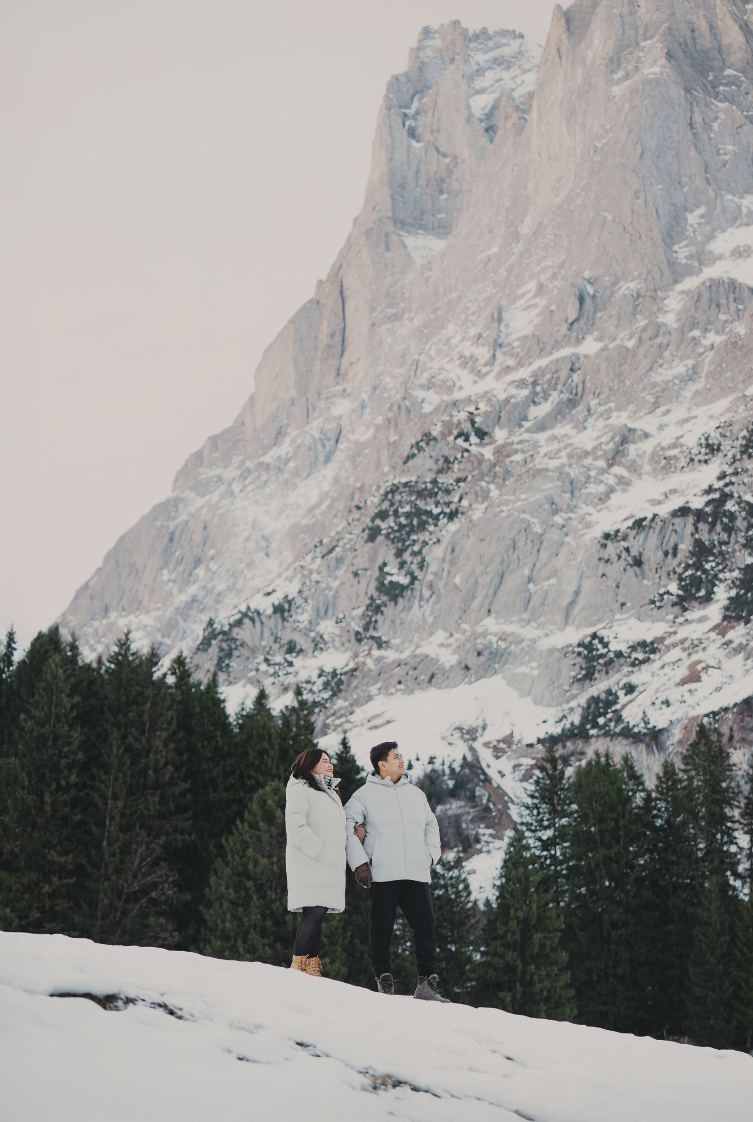Couple in matching white coats standing on snowy alpine landscape with dramatic rocky mountain backdrop.