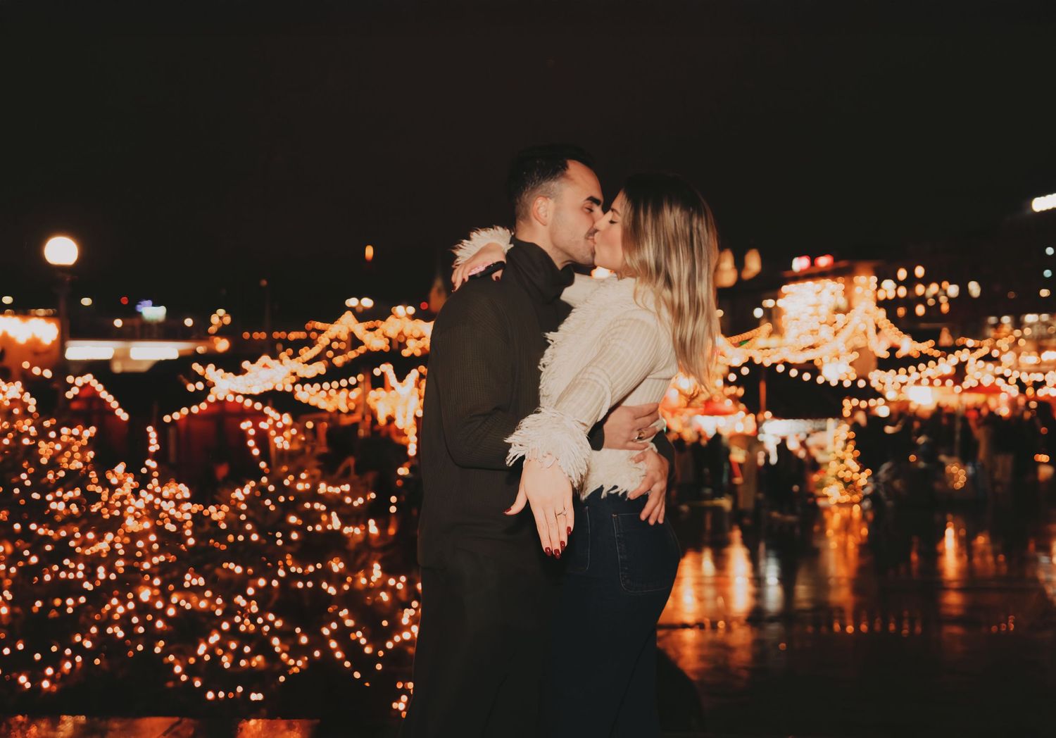 Couple kissing at a Christmas market surrounded by warm glowing holiday lights and decorations at night.