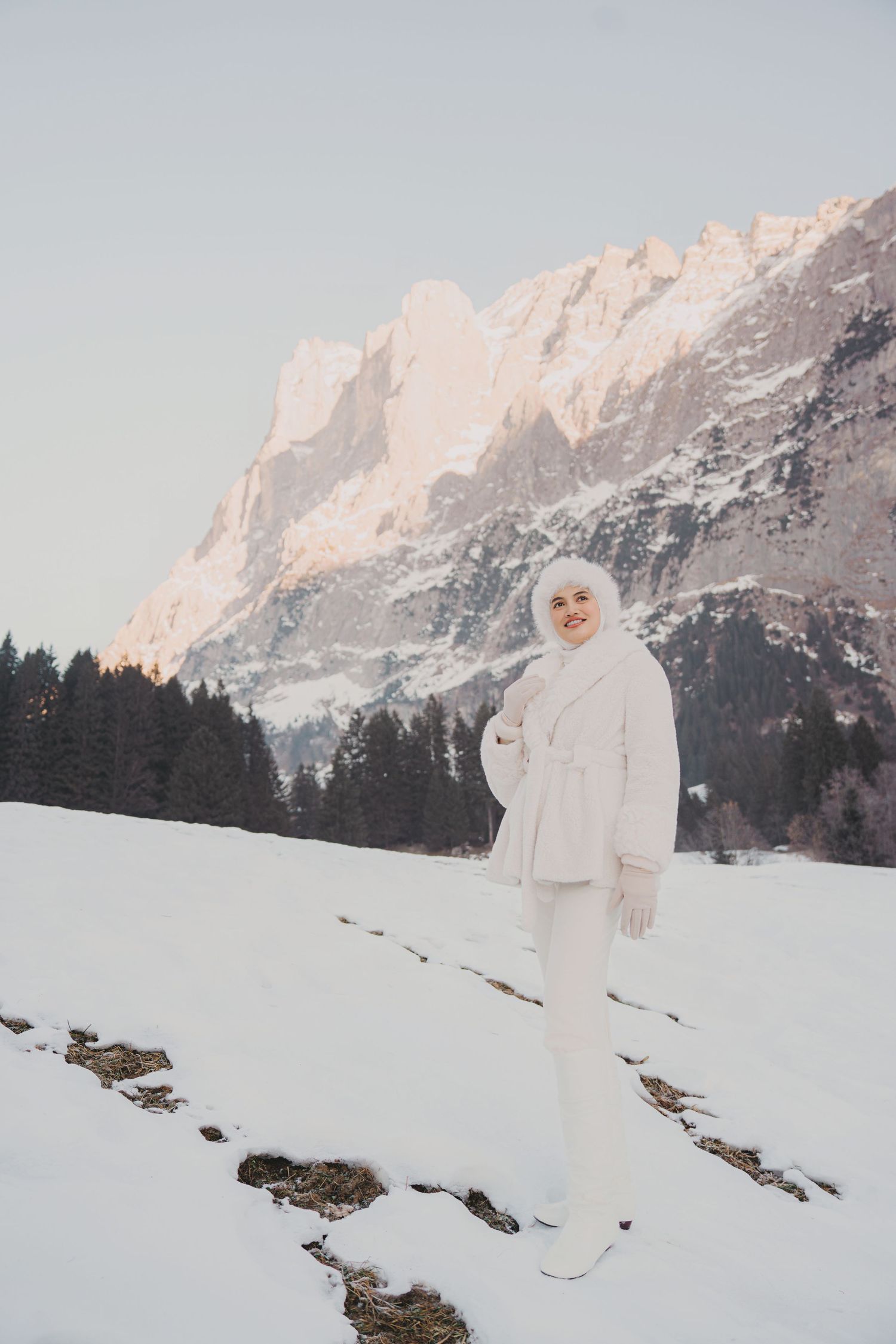 Woman in white hijab and fur coat standing in snowy alpine landscape with rocky mountain peaks behind her.