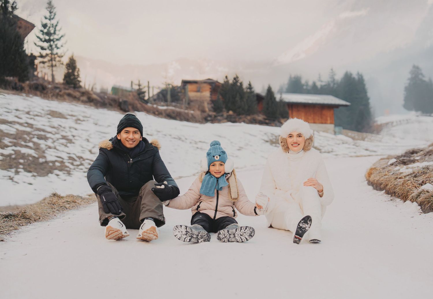 Family of three sitting together on snowy Alpine path with misty mountains and wooden chalet behind.