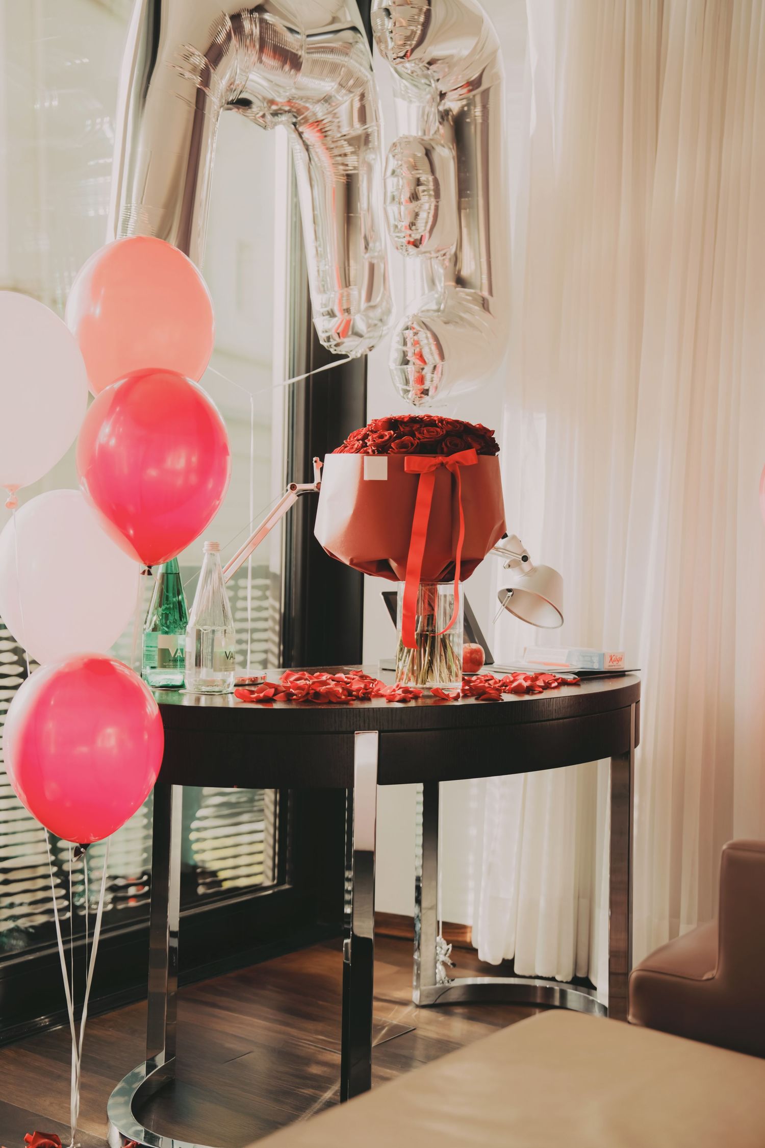 Romantic proposal setup with pink balloons, red roses in box, and silver number balloons by window.