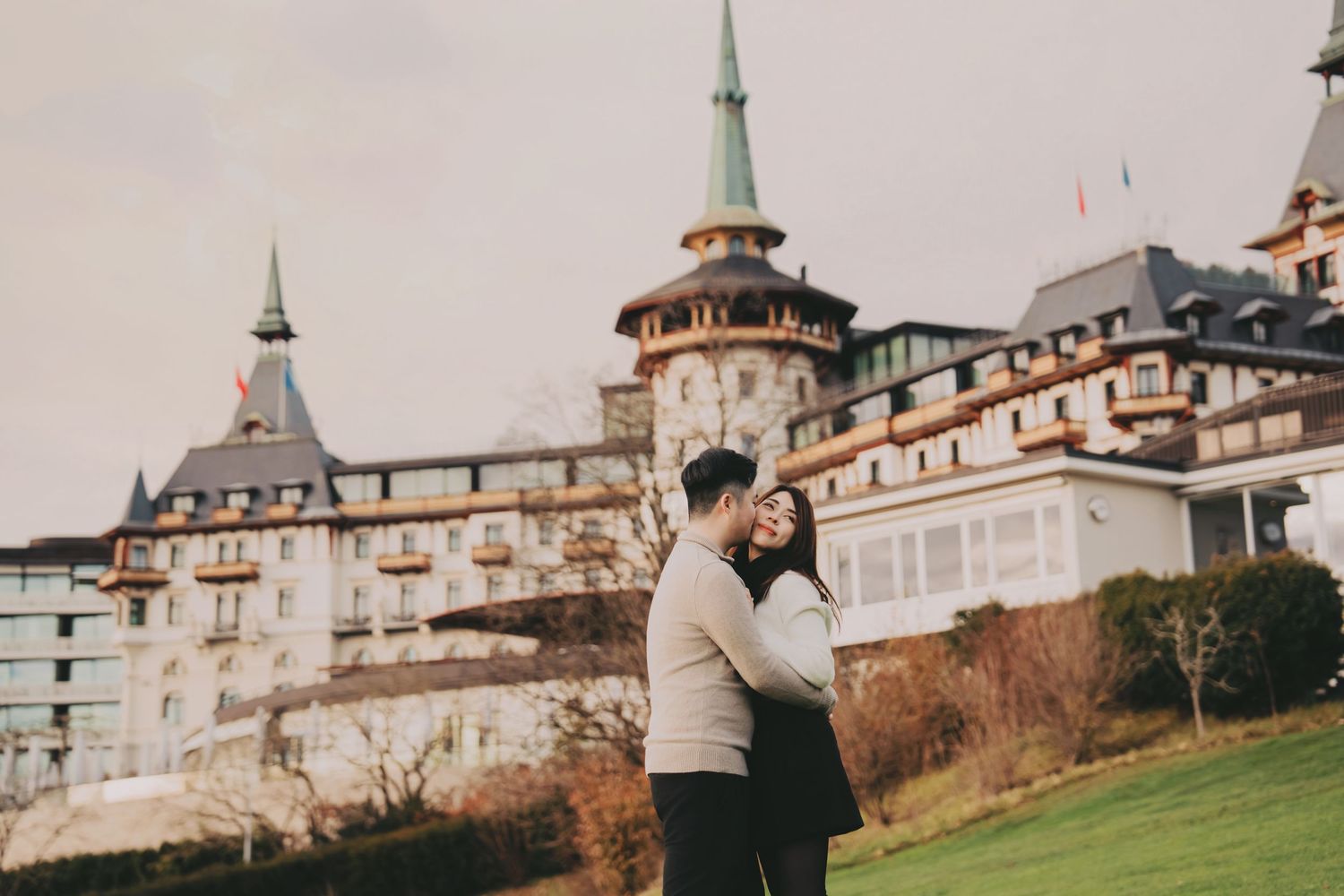 Happy couple embracing and laughing in front of a grand historic Swiss castle on a cloudy day.