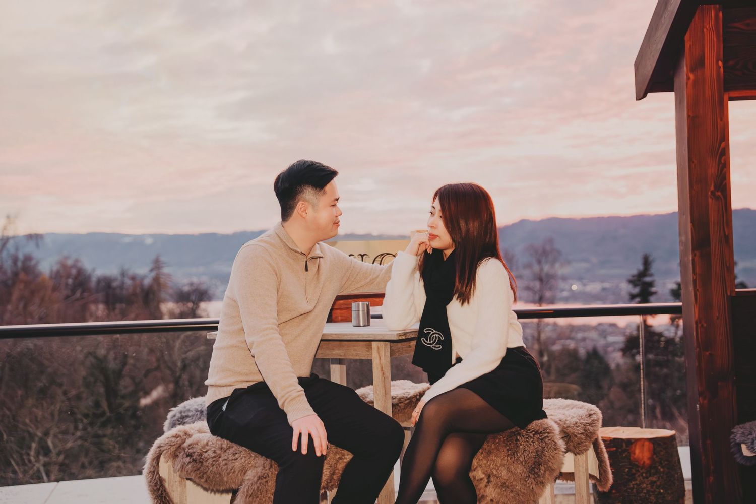 Couple sitting on fur stools on a scenic mountain terrace at sunset, sharing an intimate moment.