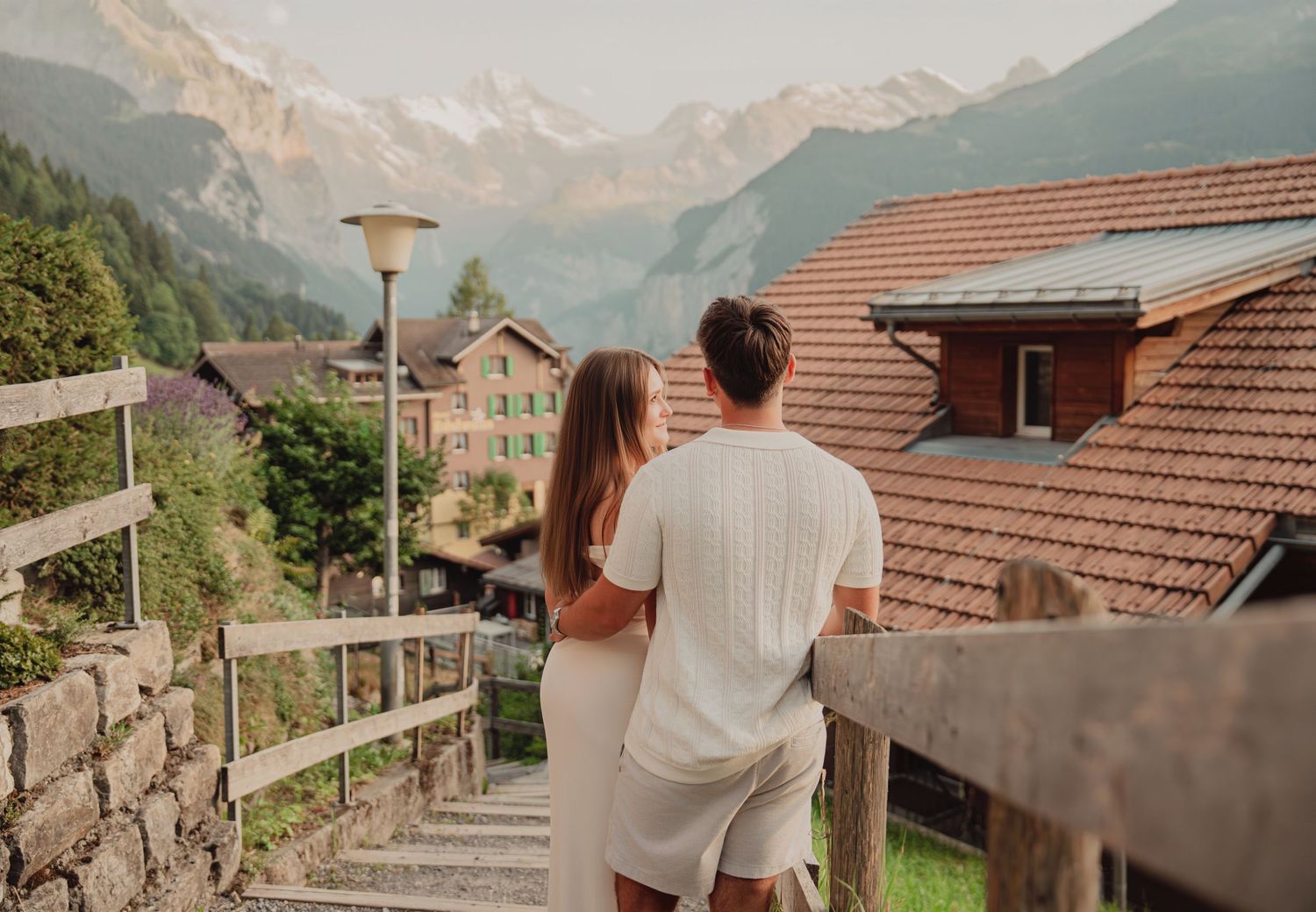 Romantic couple embracing on wooden balcony overlooking picturesque Swiss Alpine village and misty mountain peaks.