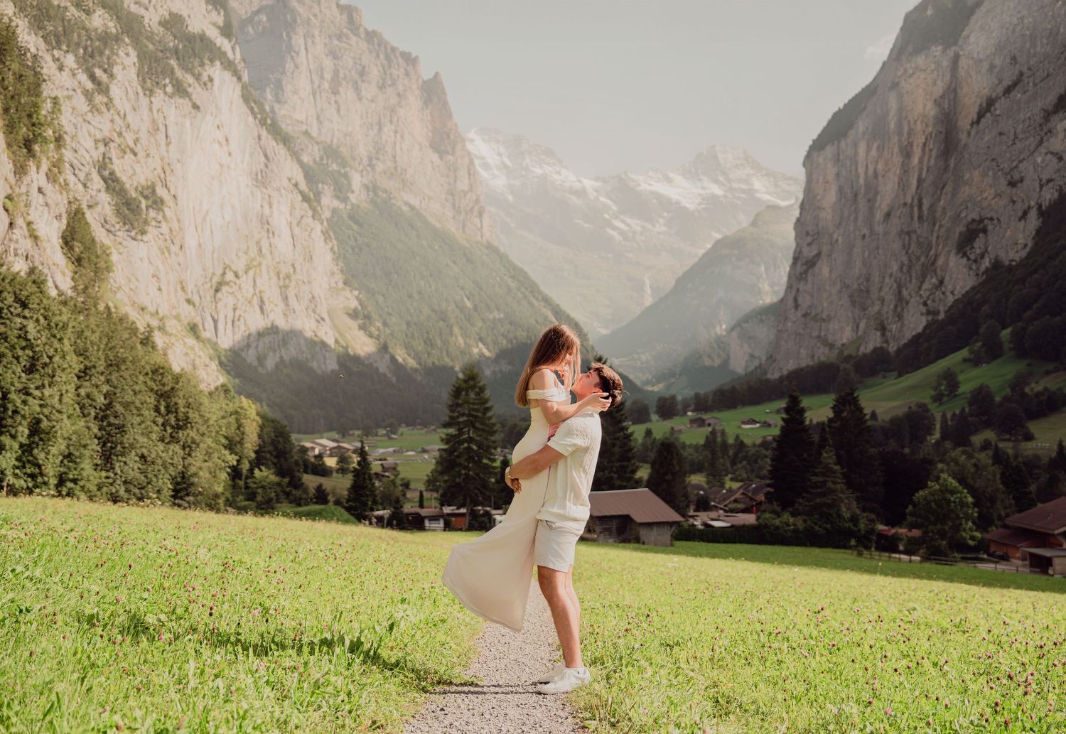 Man lifting woman in white dress on a gravel path with dramatic Swiss Alps valley in background.
