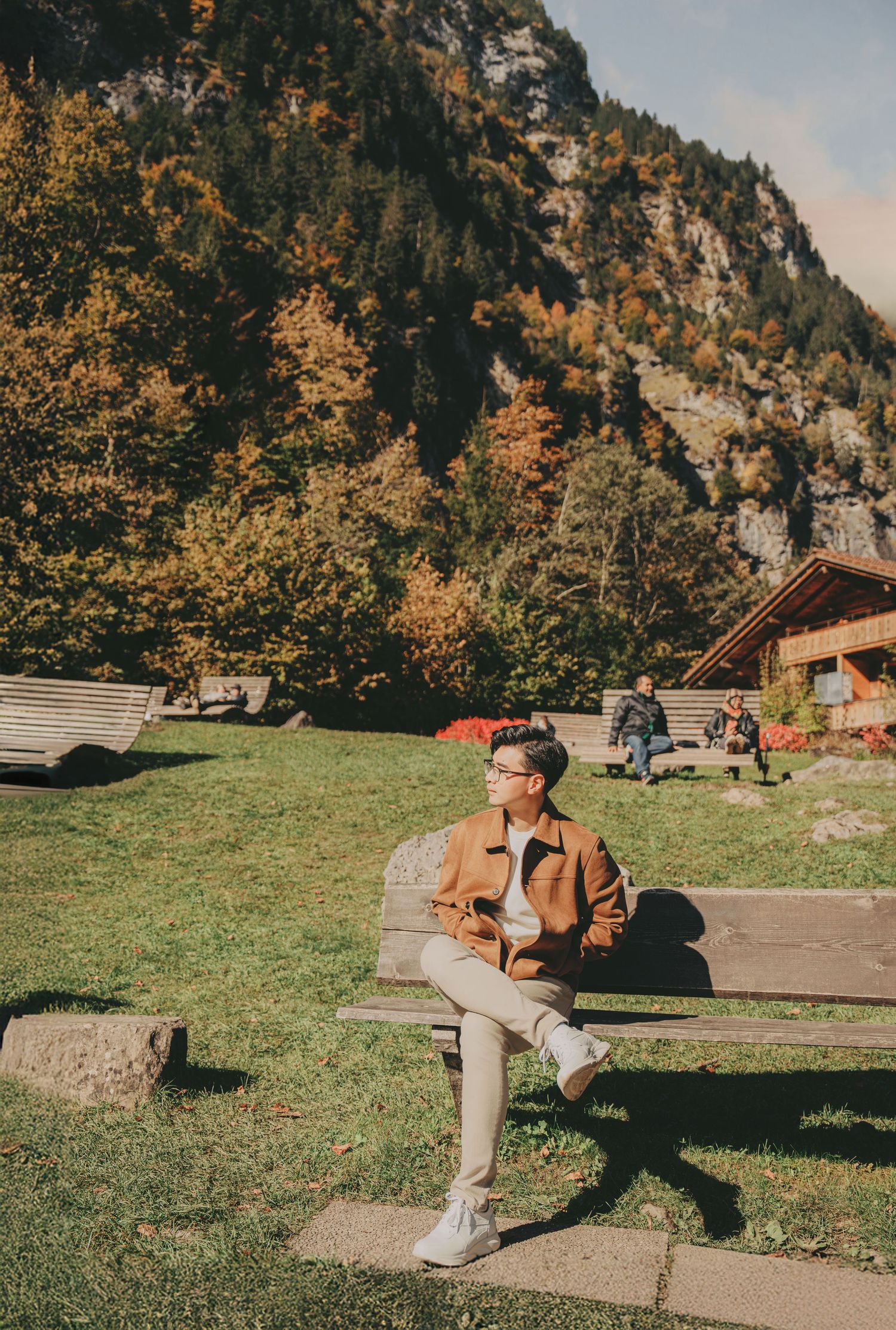 Young man in brown jacket sitting on bench in colorful autumn mountain landscape.