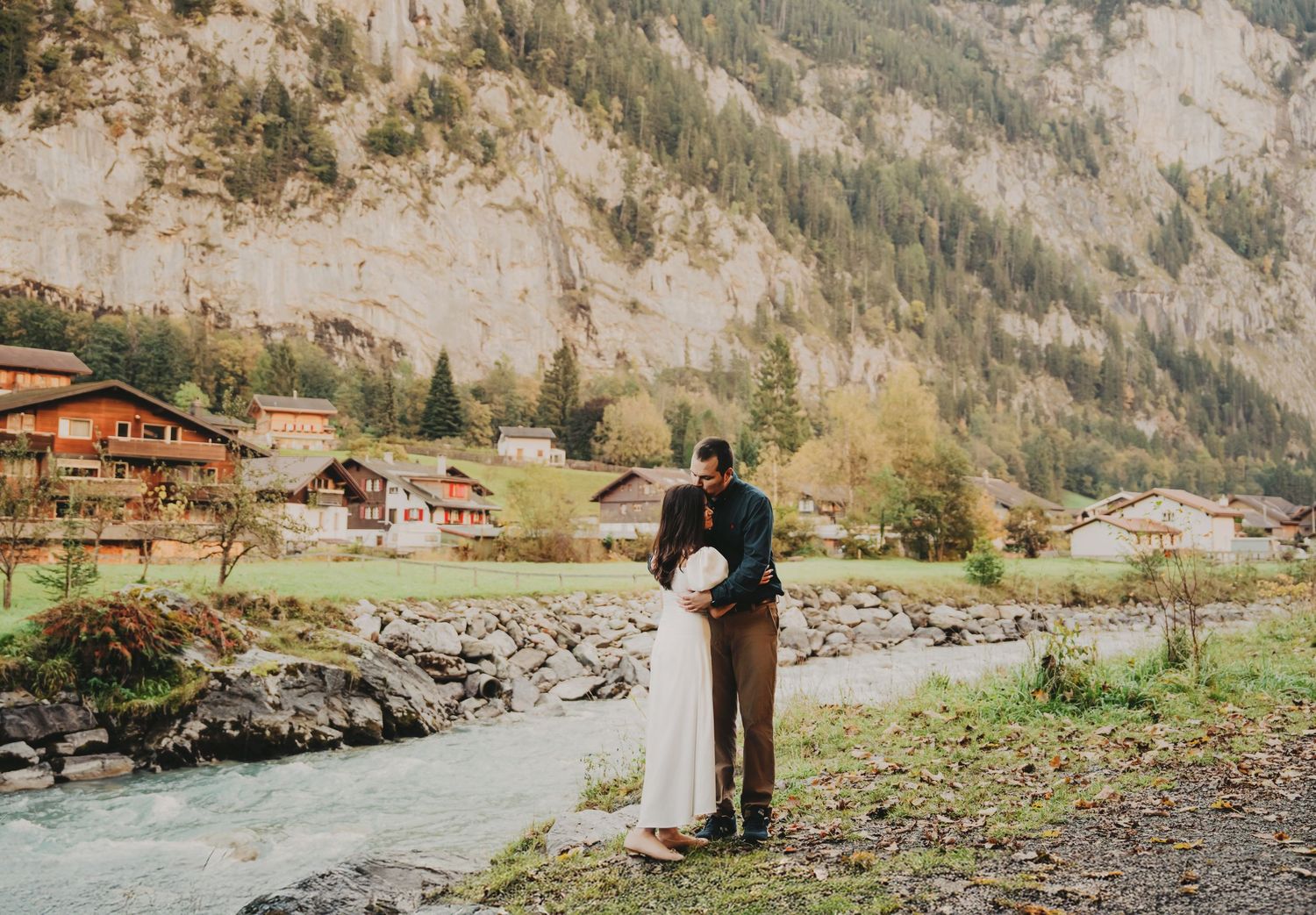 Couple embracing by a rocky river with Swiss mountain village and cliffs behind them.
