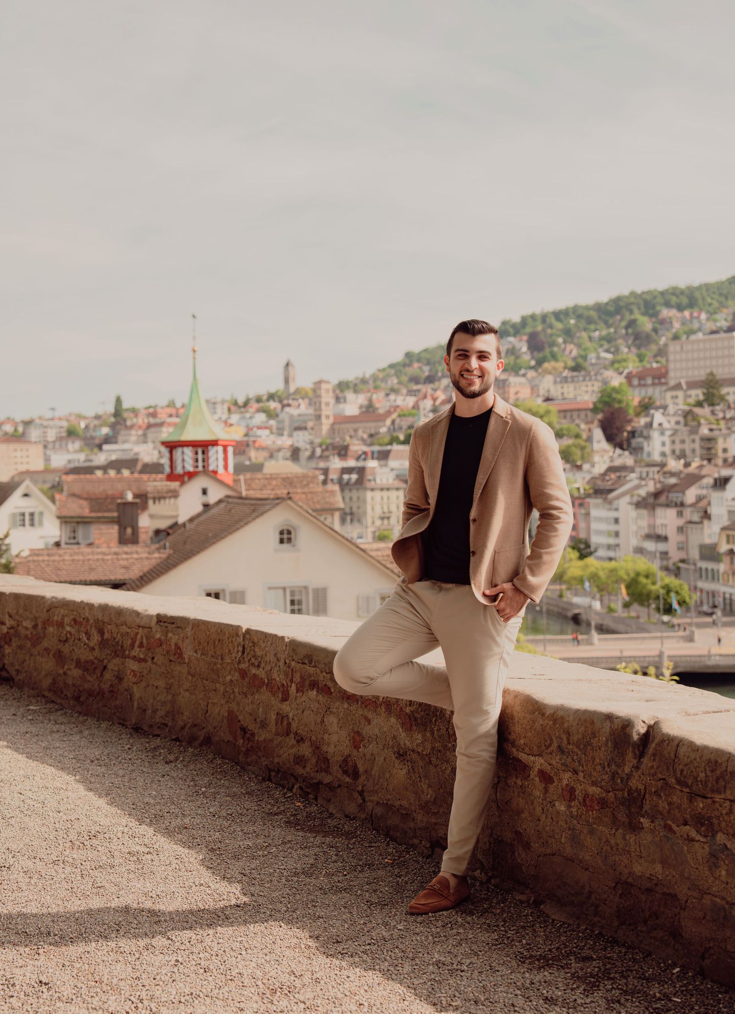 Stylish man in beige blazer and black shirt leans against stone wall with Zurich cityscape behind him.