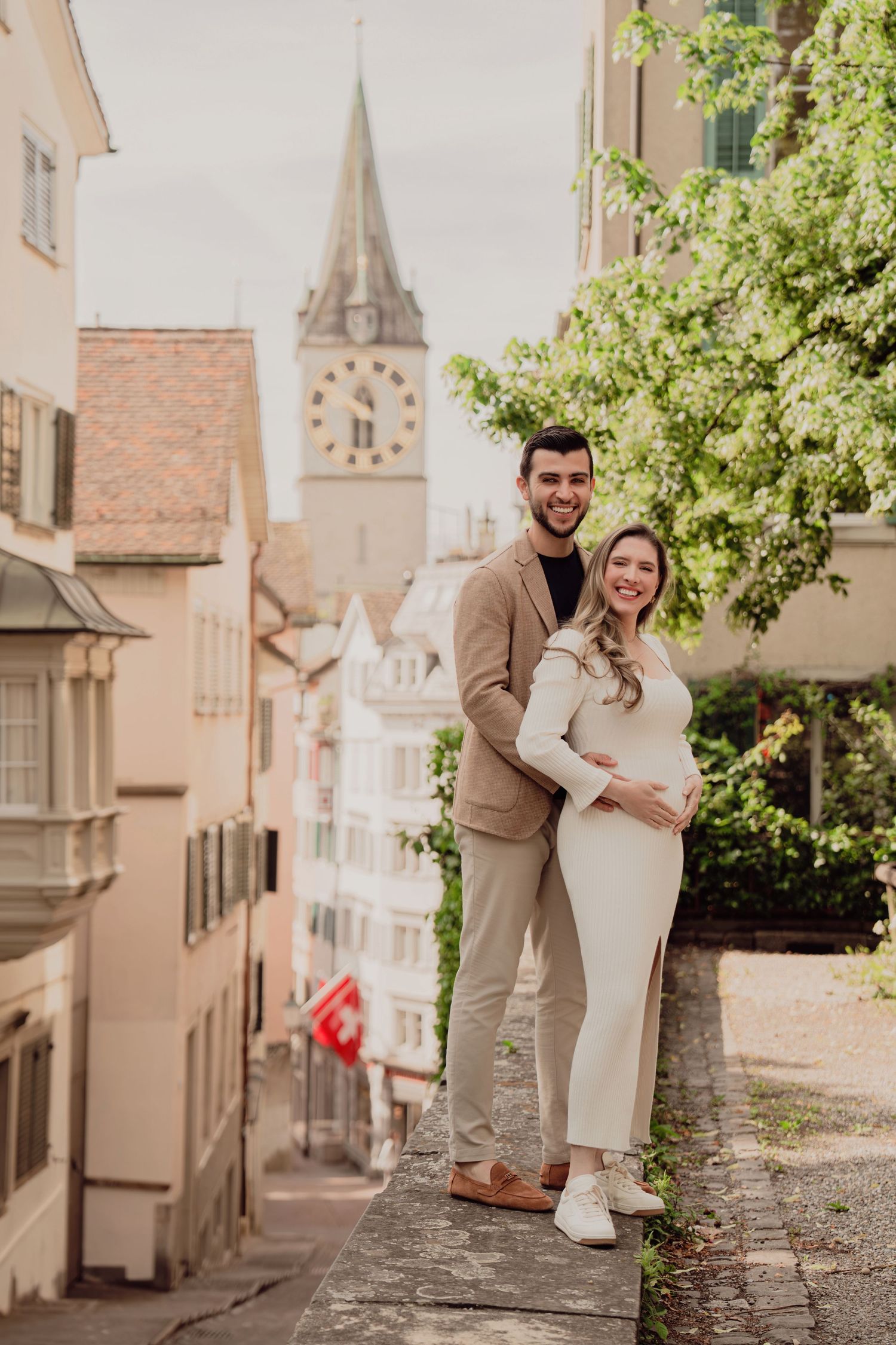 Happy pregnant couple posing on a terrace with a European clock tower and Swiss flag in the background.