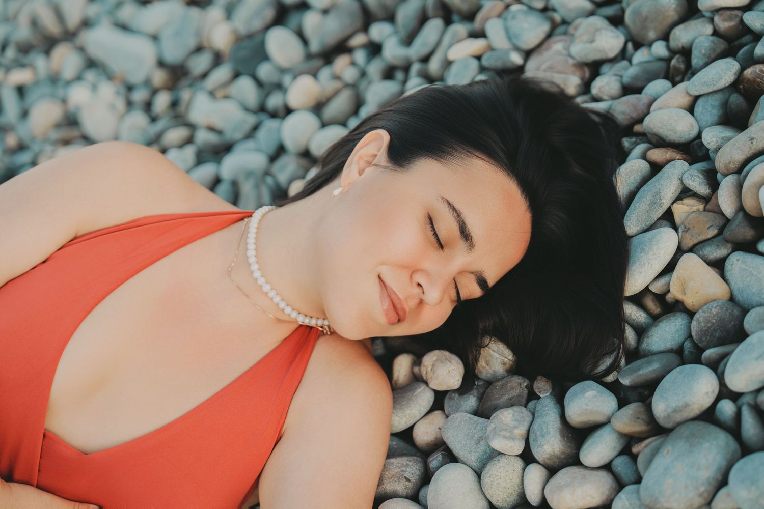 Woman in red swimsuit and pearl necklace lying on pebble beach with eyes closed.