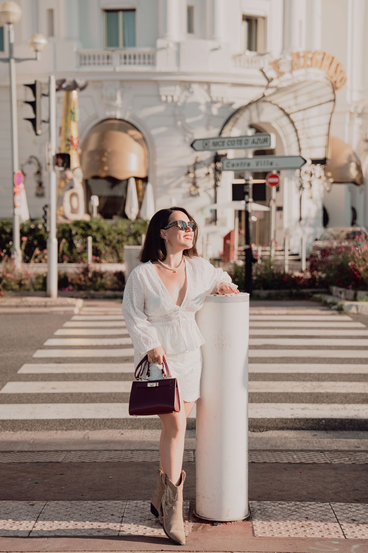 Stylish woman in white ensemble with burgundy handbag posing near crosswalk in Nice.