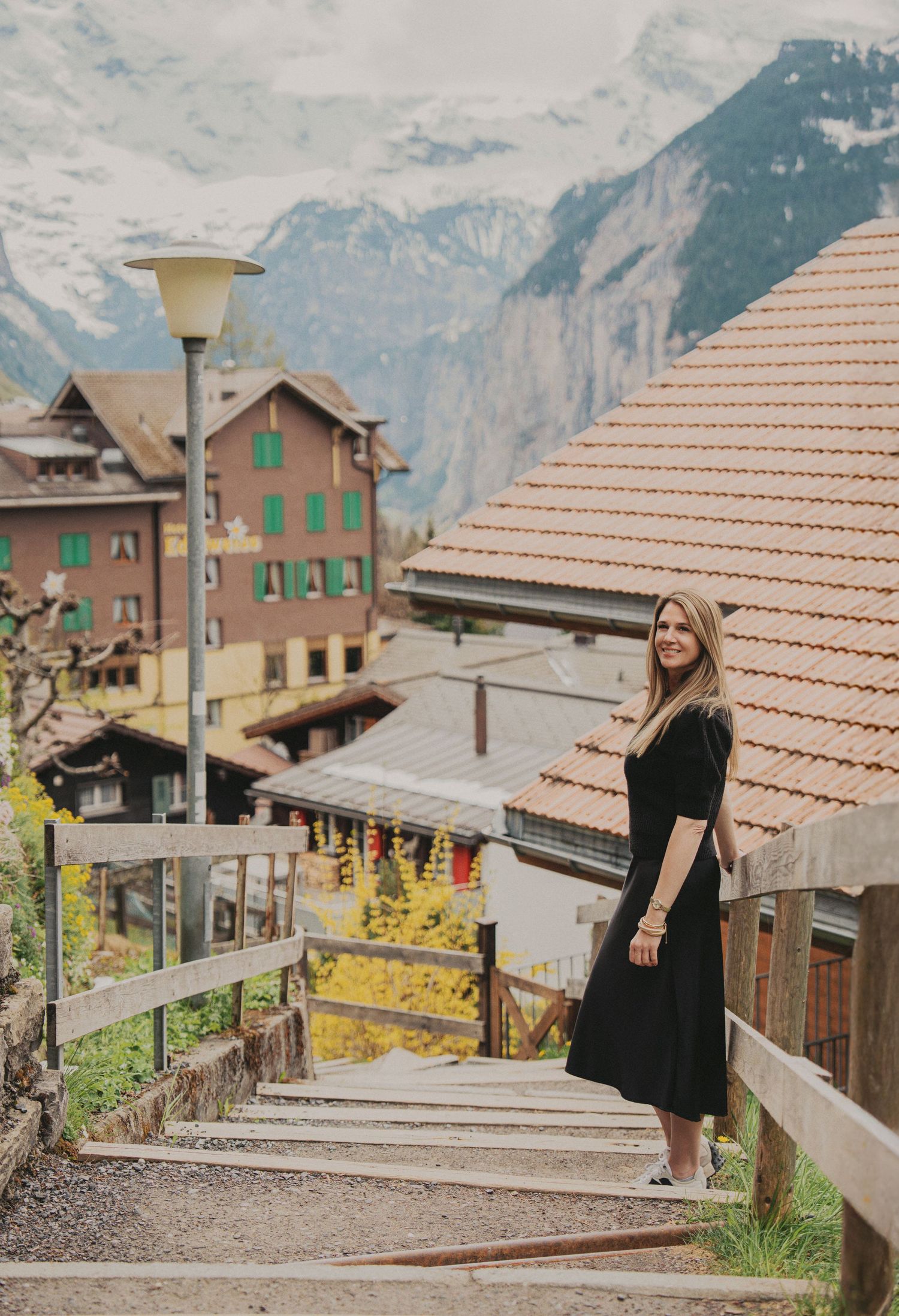 Woman in black dress standing on scenic stairway in Swiss Alpine village with snow-capped mountains behind.