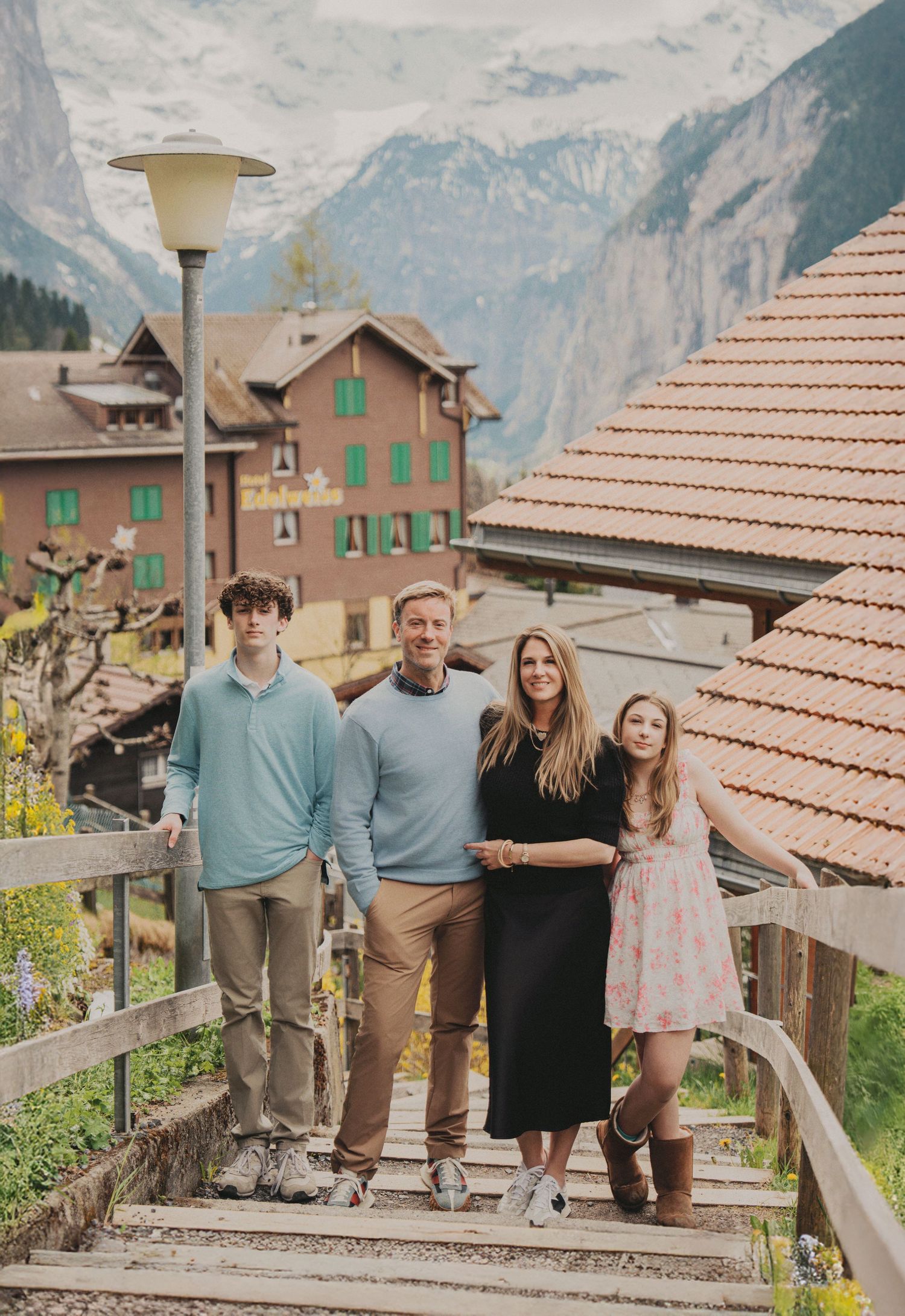 Family of four posing on wooden steps in a Swiss Alpine village with snow-capped mountains backdrop.