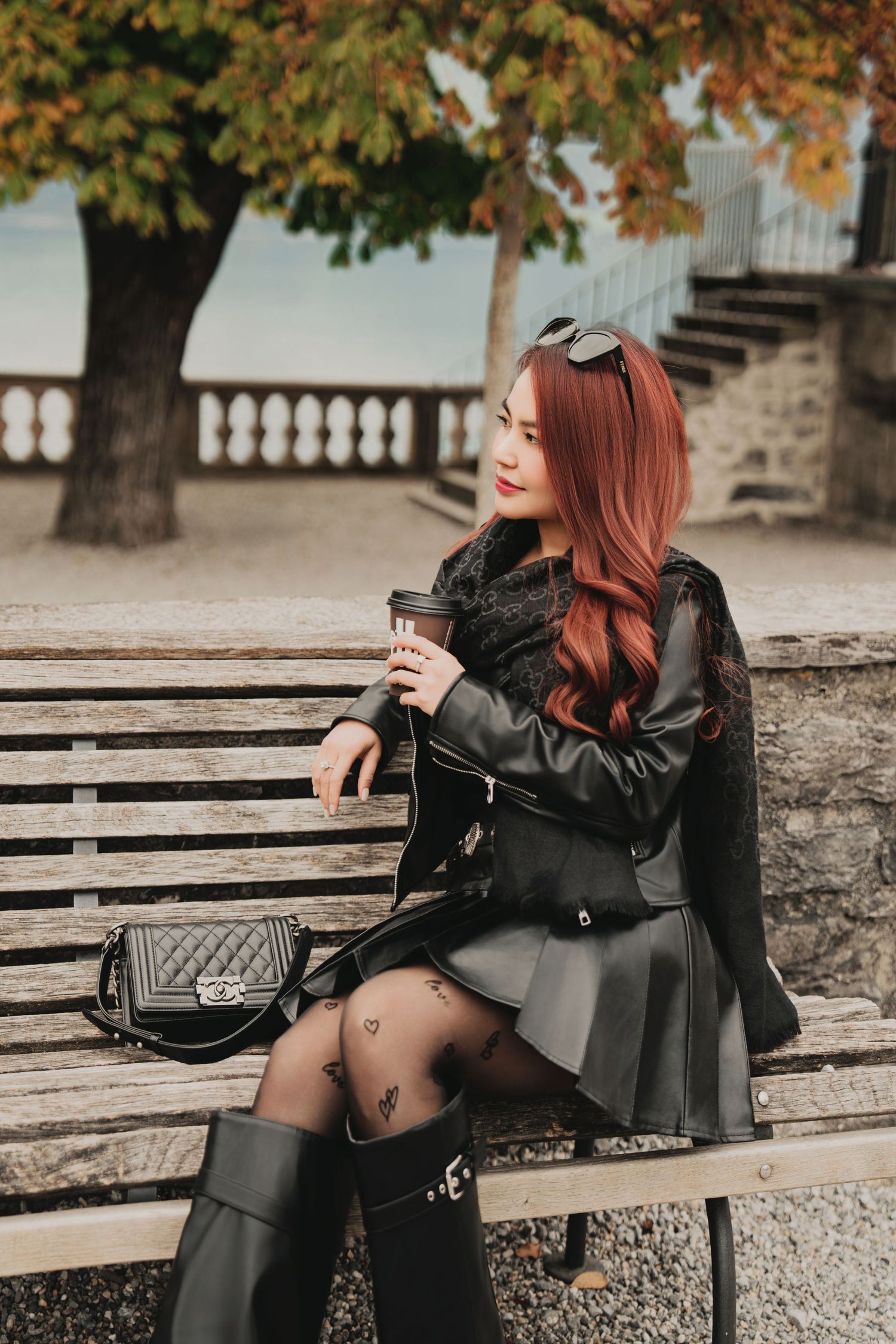 Fashionable red-haired woman in black leather skirt sits on bench holding coffee cup in autumn park.