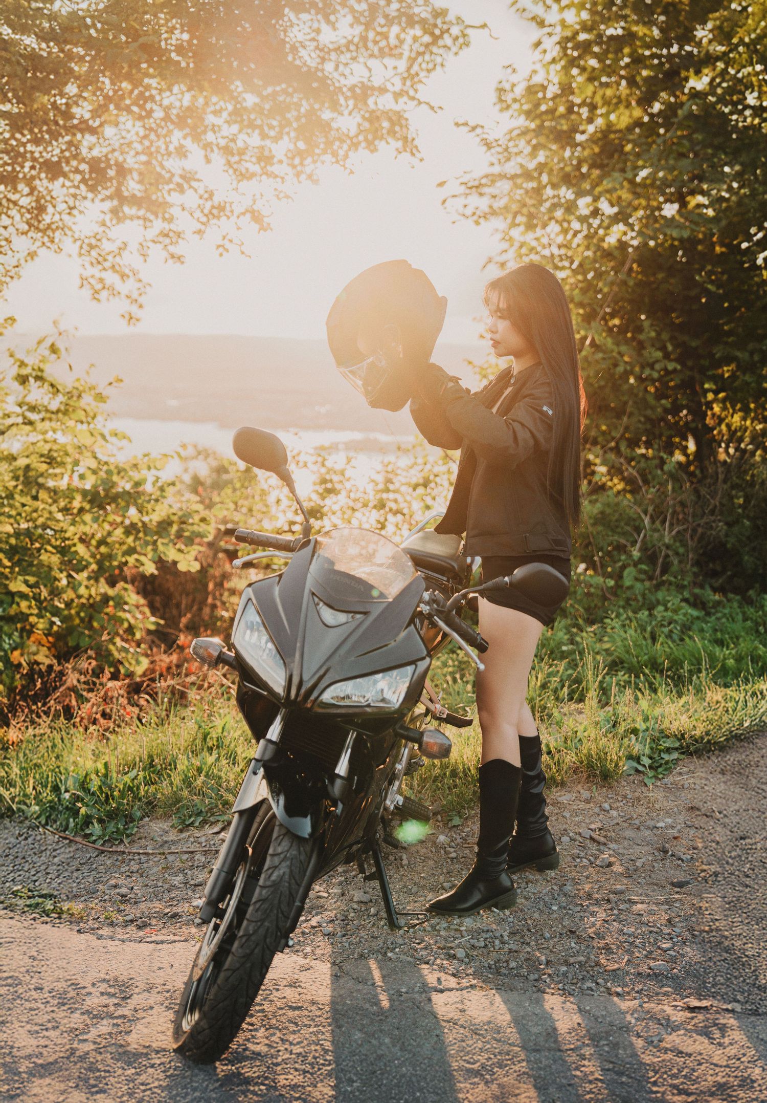 Woman holding a motorcycle helmet beside a sport bike on a sunlit road surrounded by lush green trees.