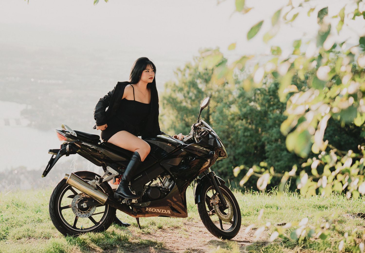 Young woman in black outfit posing on a Honda motorcycle on a lush hillside with misty river backdrop.