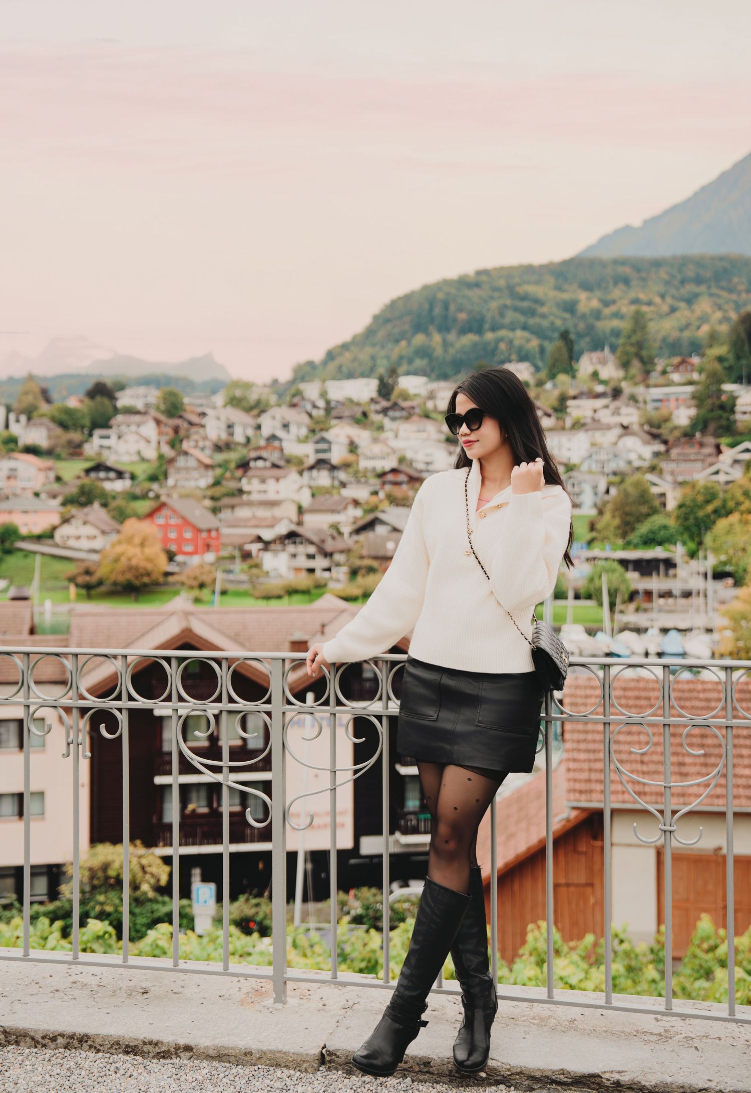 Stylish woman in cream sweater and leather skirt stands by ornate railing with European town backdrop.