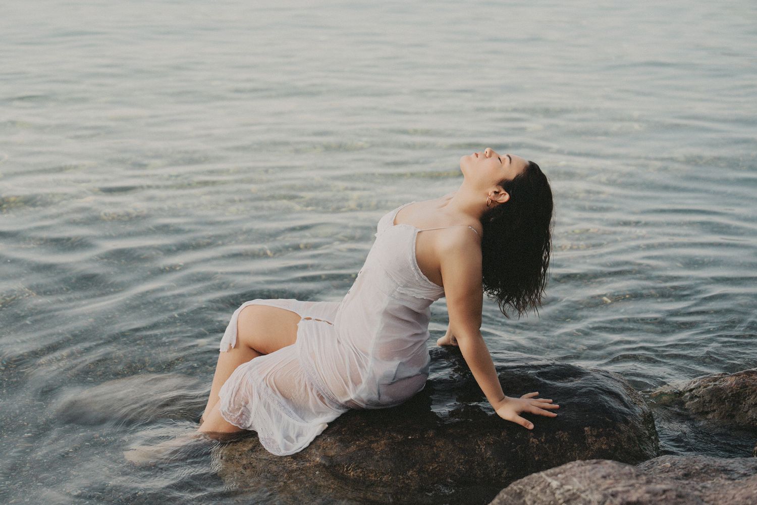 Woman in wet white dress sitting on rocks by calm water, head tilted back in serene pose.