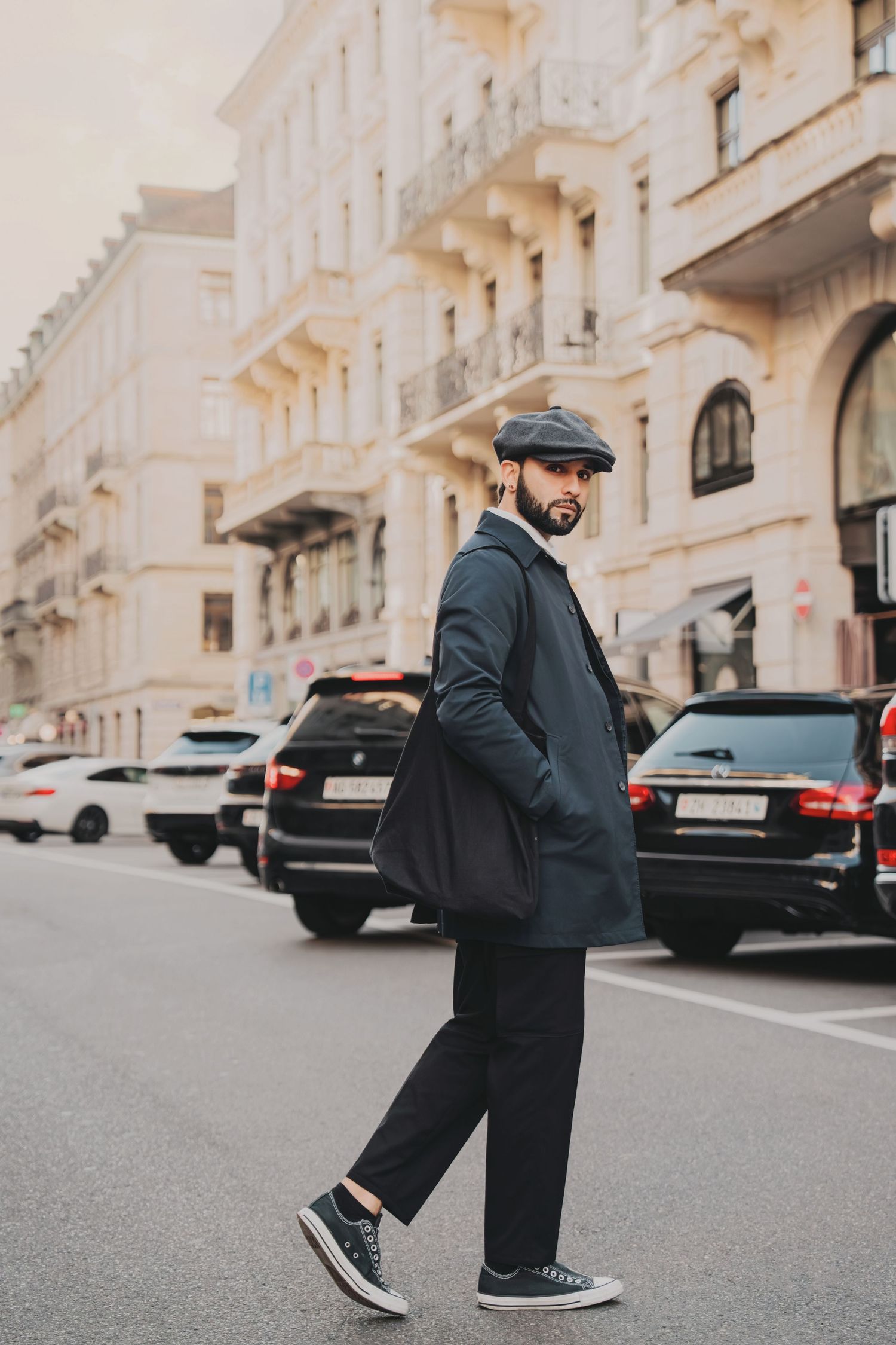 Bearded man in dark coat and flat cap crossing cobblestone street with luxury cars and classical buildings.