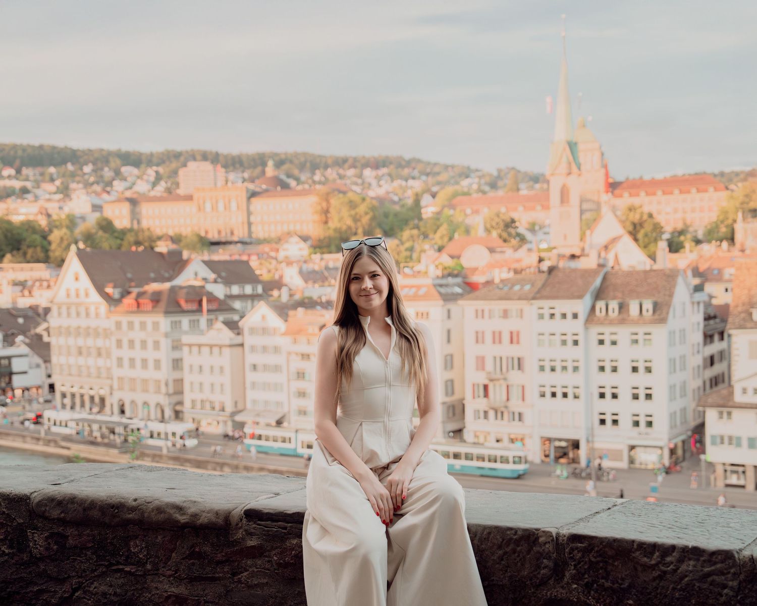 Woman in white dress sits on stone wall with panoramic view of Zurich cityscape behind her.