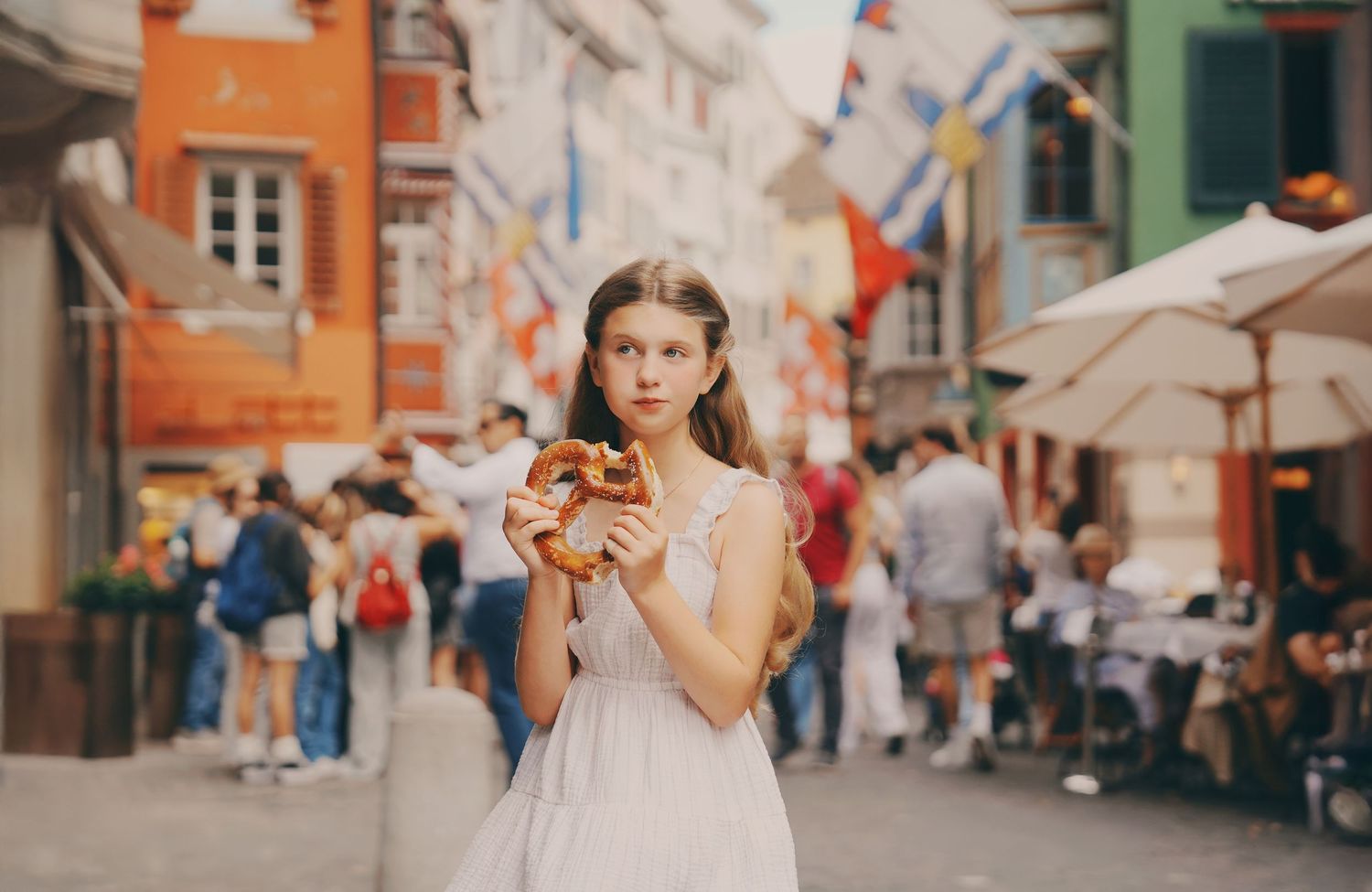 A young girl in a white dress holds a large pretzel on a busy European street with colorful flags.
