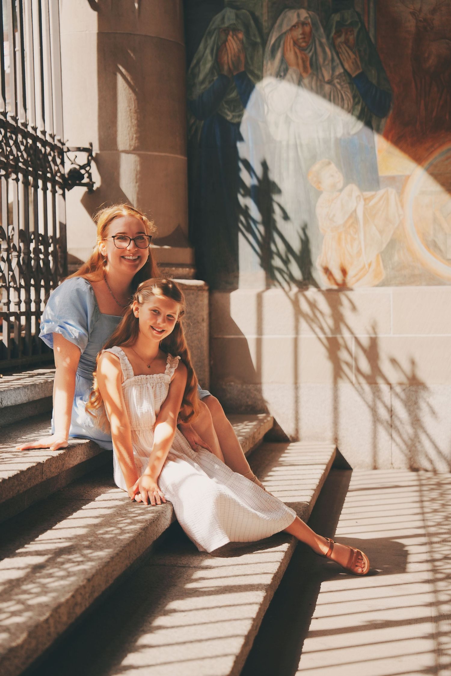 Mother and daughter sitting on sun-dappled church steps with religious fresco mural backdrop.