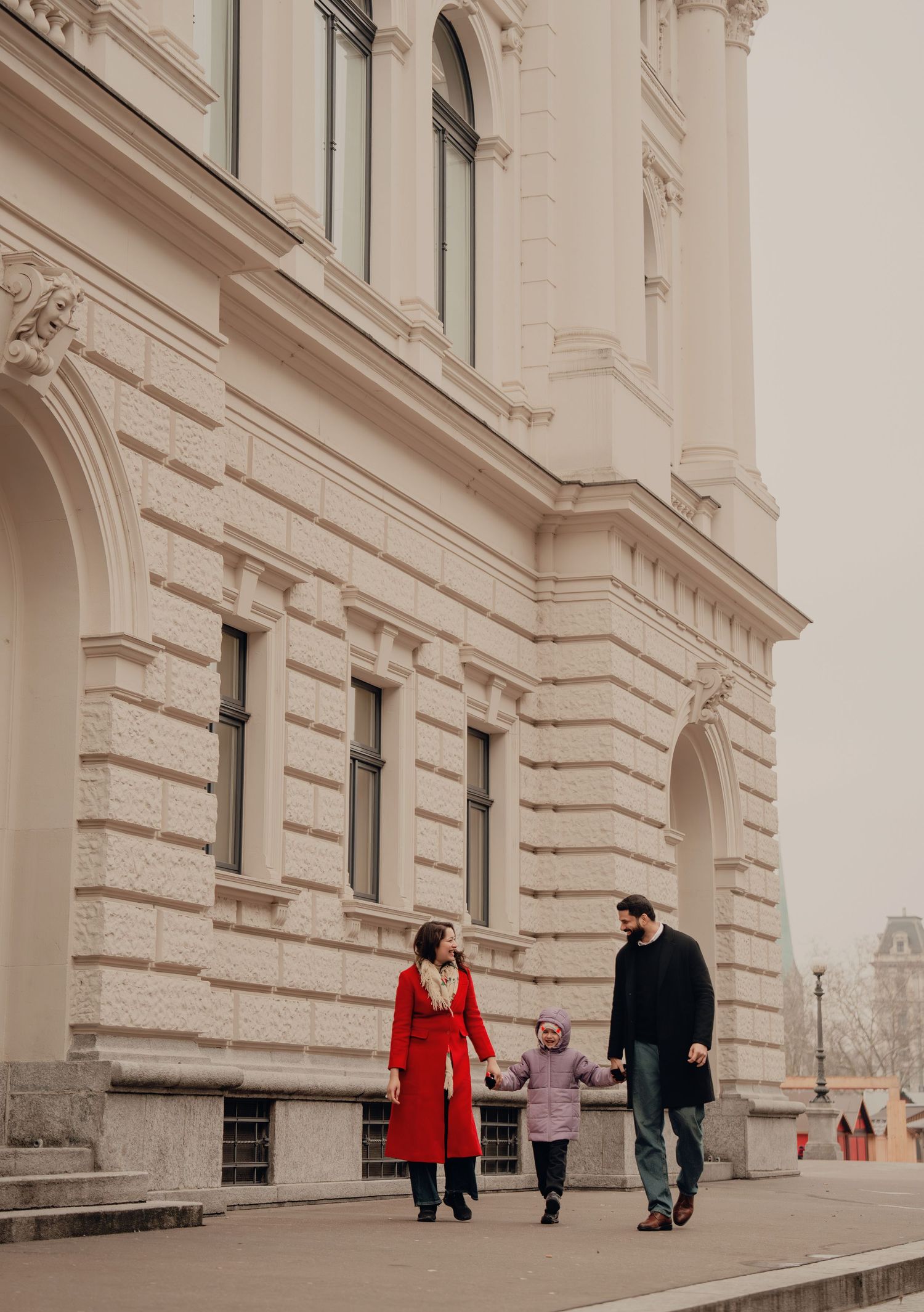 Happy family of three walking hand in hand past a grand neoclassical building facade.