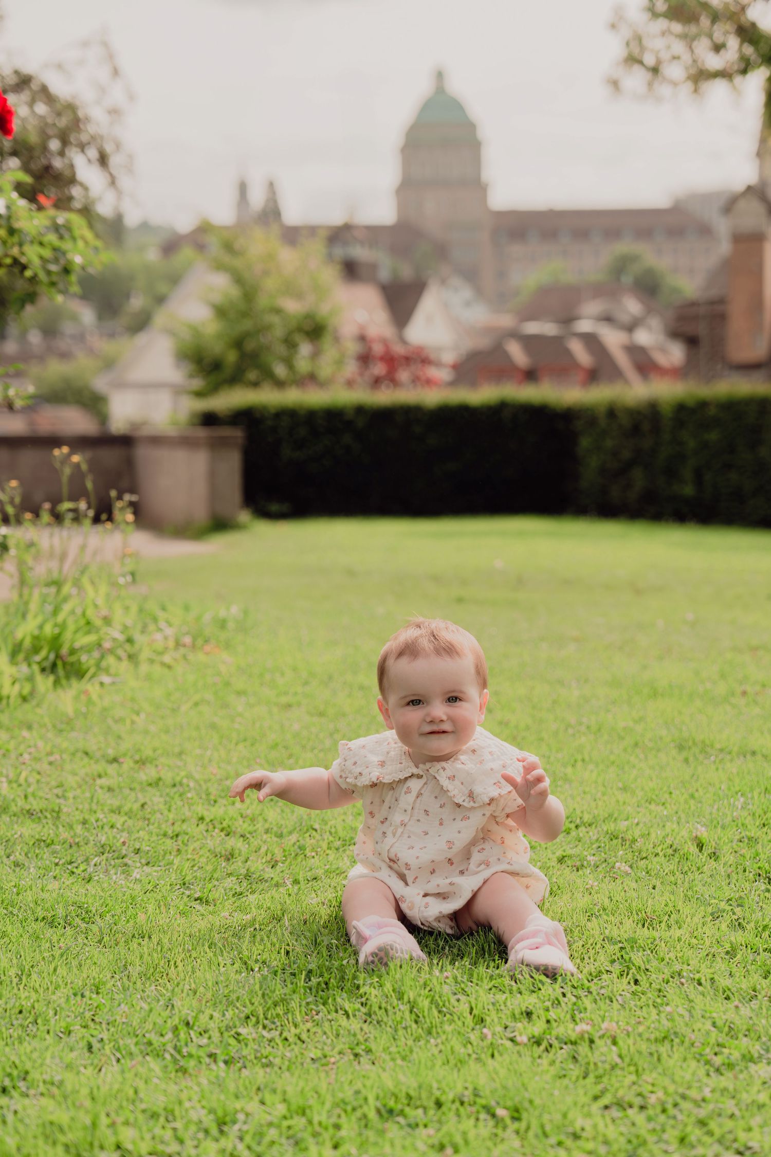 Adorable baby in floral romper sitting on green grass with European city skyline in background.