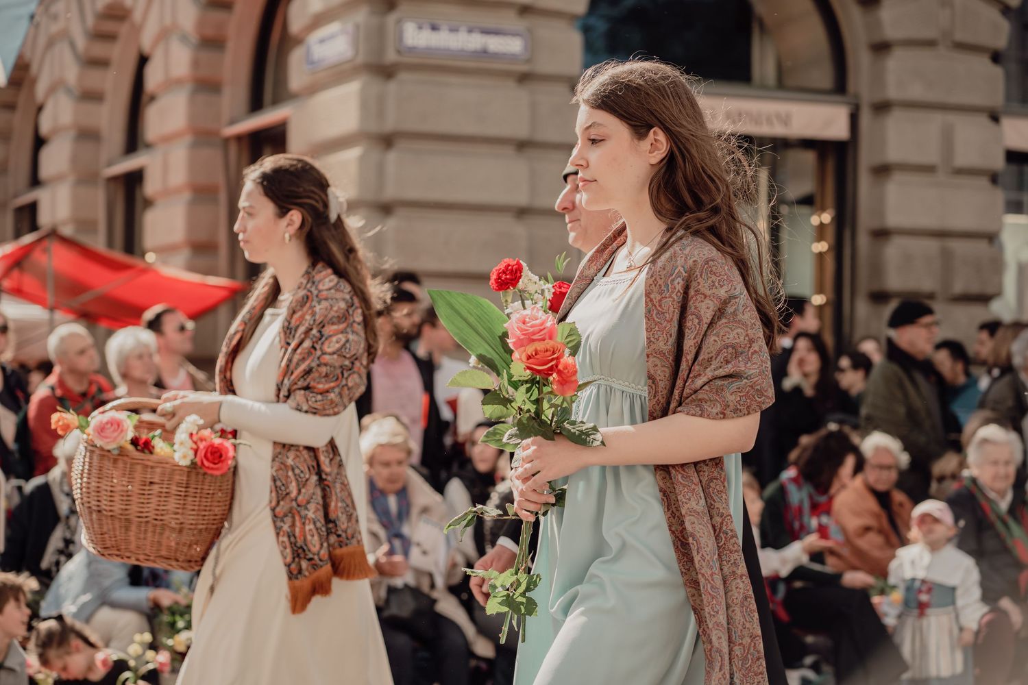 Young women in vintage dresses carry roses and flower baskets in a festive street parade.