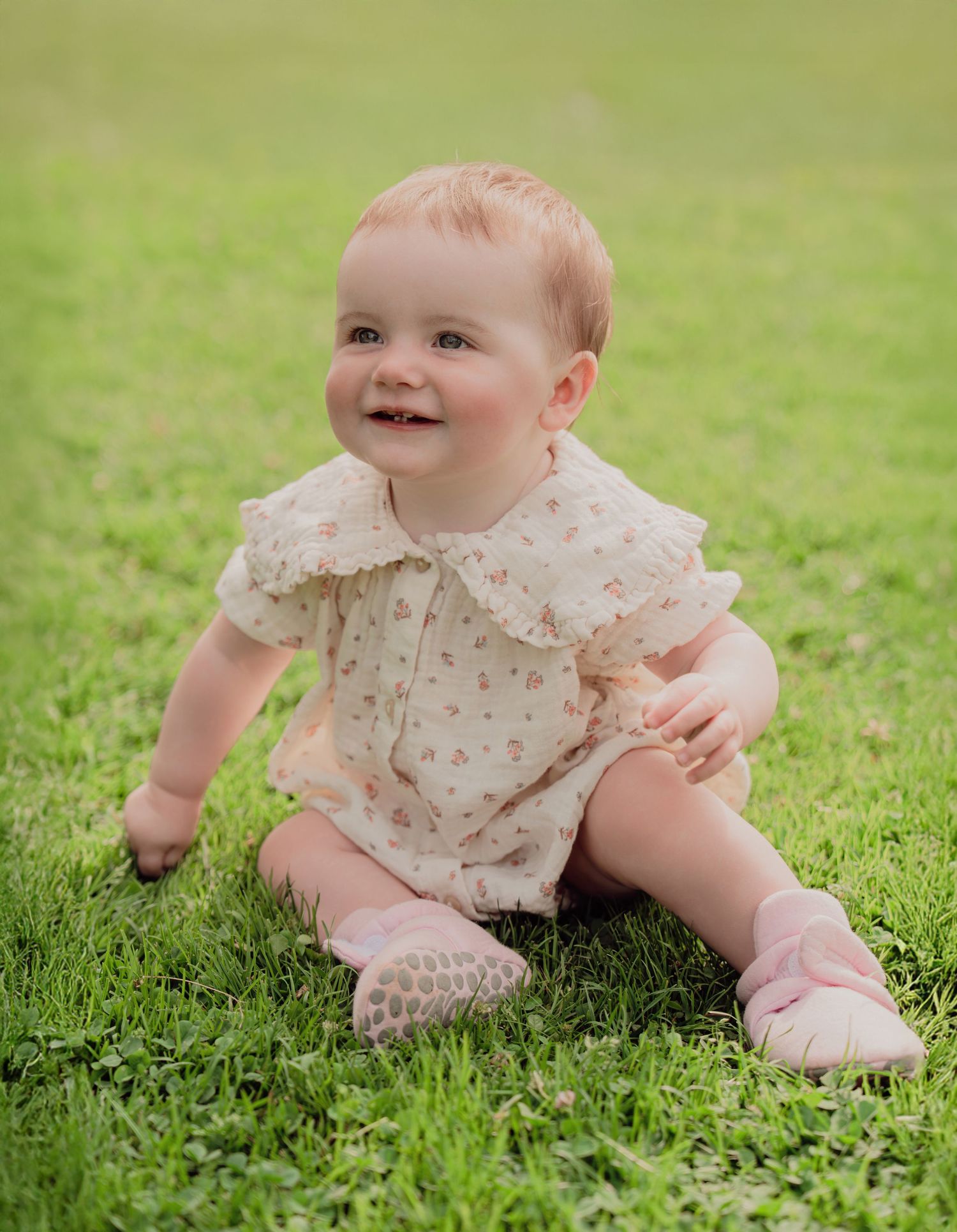 Smiling baby in cream floral romper with pink socks sitting on lush green garden grass.